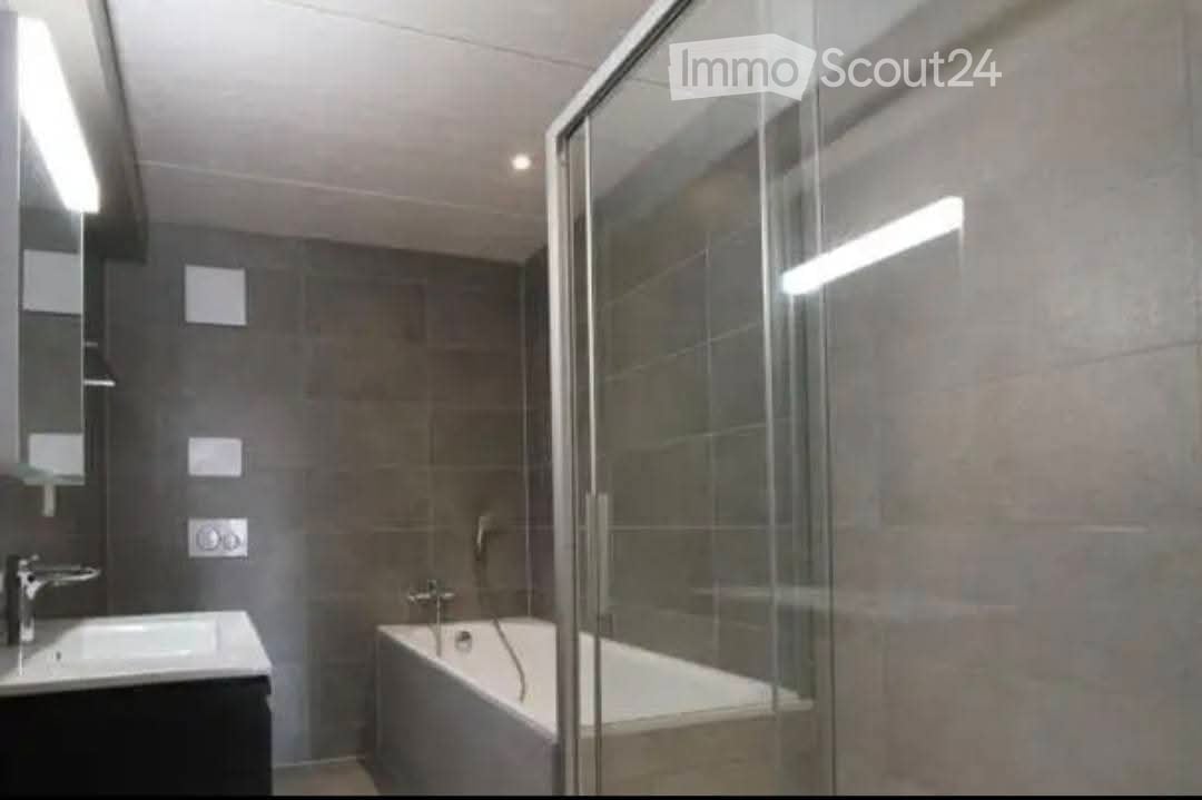 Modern bathroom with a bathtub, sink, and glass shower enclosure. The walls are tiled in a neutral color, and the lighting is bright.