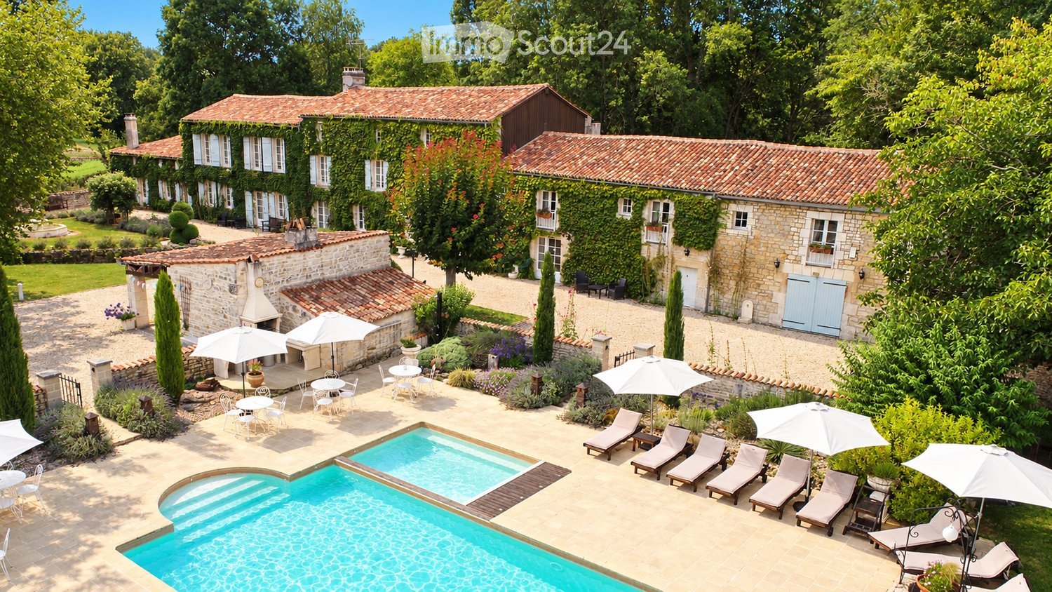 Aerial view of a large, rustic stone manor surrounded by lush greenery, featuring a swimming pool, outdoor seating areas with tables and chairs, sun loungers, and umbrellas.