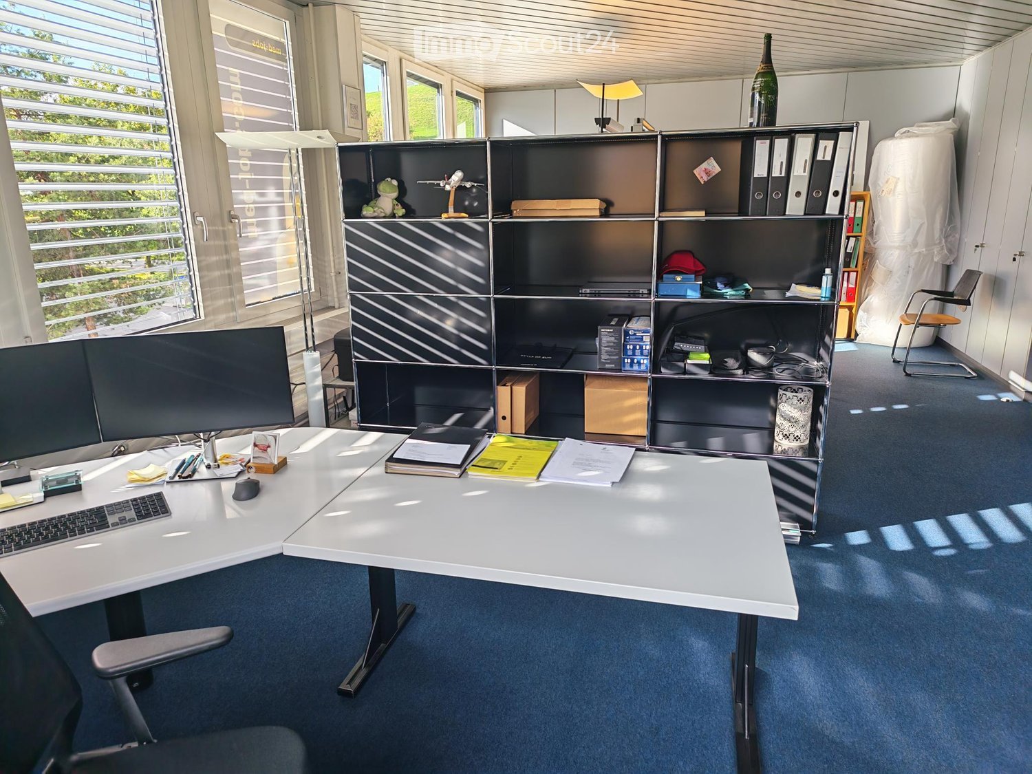 Modern office space with a white desk, ergonomic chair, large screen, keyboard, mouse, and a black shelving unit holding various items including a lamp, bottle, and decorative objects.