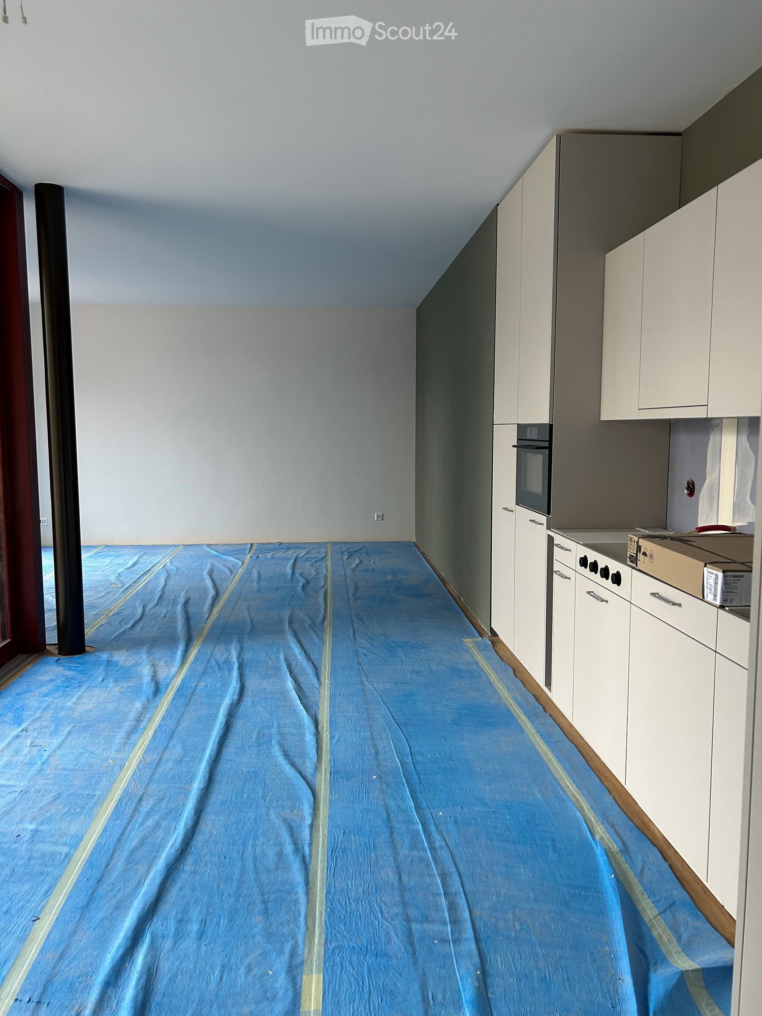 Empty room with white walls, a blue protective floor covering, and white kitchen cabinets along one wall.