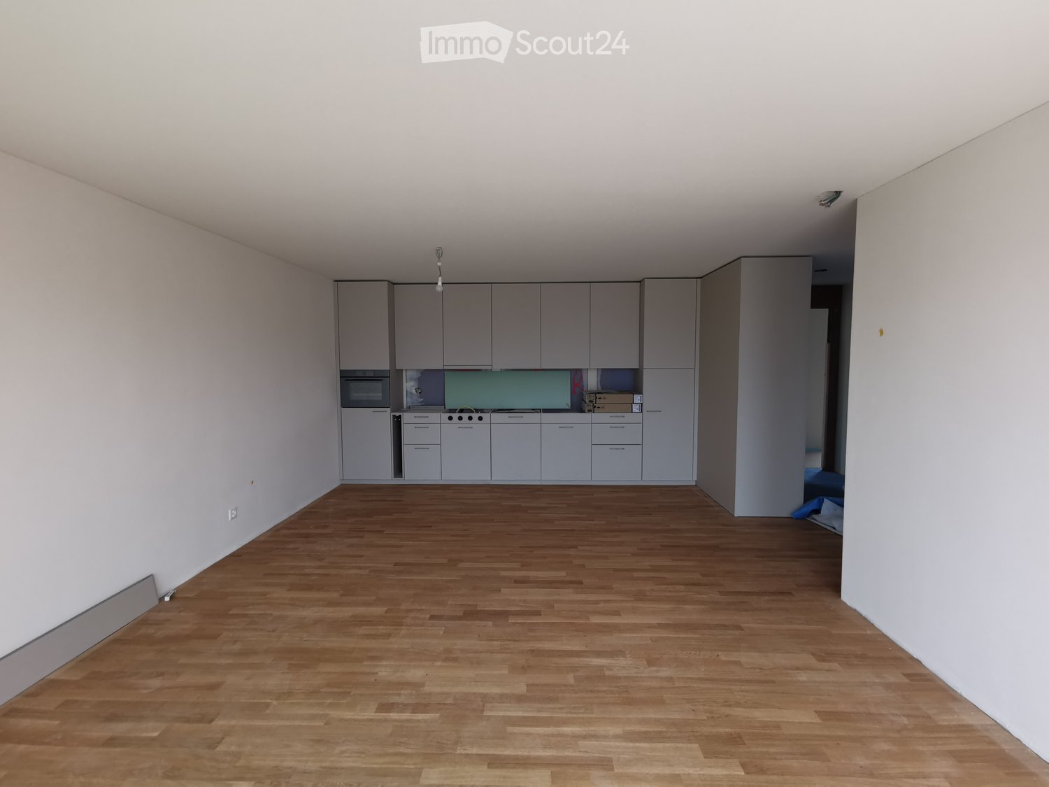 Empty room with light wooden flooring and white walls, featuring a kitchen area with cabinets and appliances in the background.