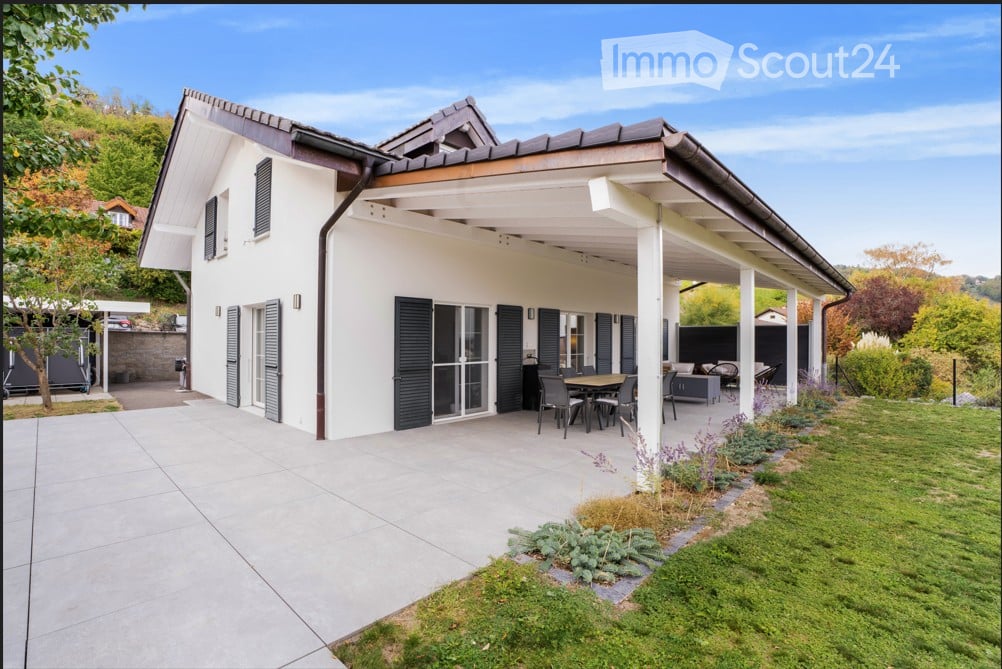 Modern two-story house with white exterior, dark shutters, and a covered patio area with outdoor furniture.