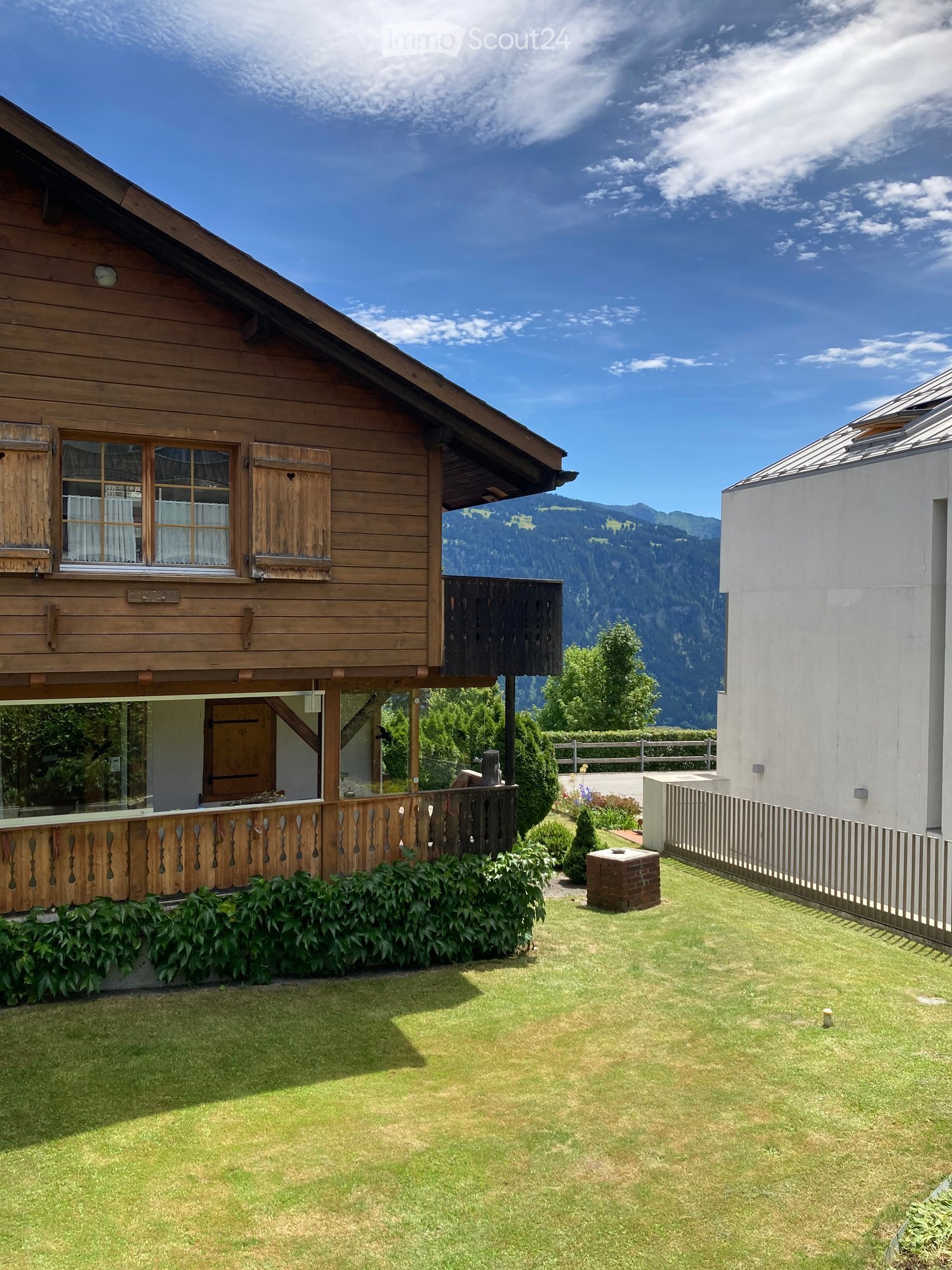 A two-story wooden house with a balcony, surrounded by a well-maintained lawn and garden, with a scenic mountain view in the background.