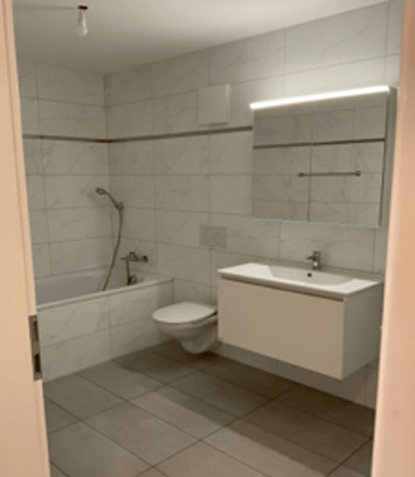 Modern bathroom with a bathtub, toilet, sink, and a large mirror with built-in lighting. The walls are tiled with a mix of light and dark tiles, and the floor is covered with beige tiles.