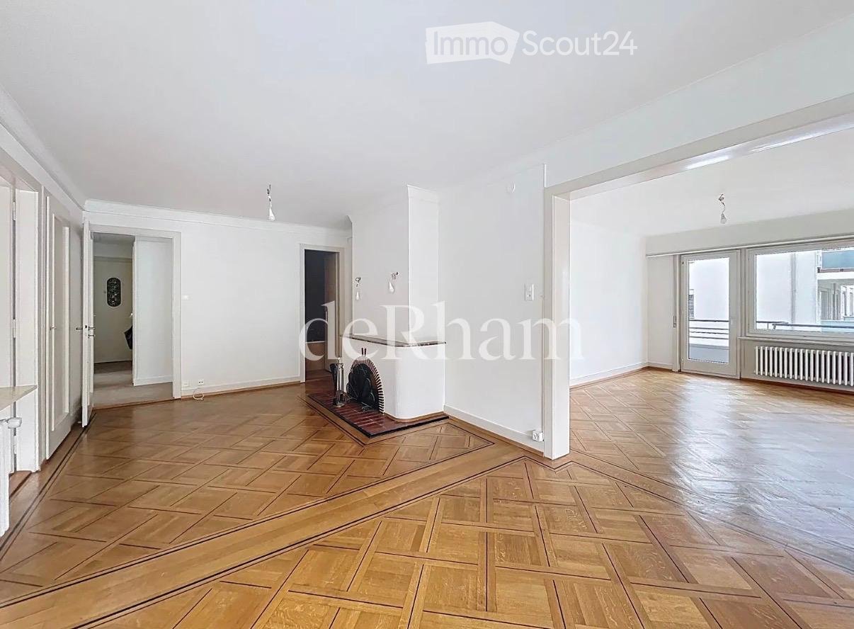 Empty room with wooden parquet flooring, white walls, and large windows. Features a fireplace with a decorative screen and a radiator under the windows.