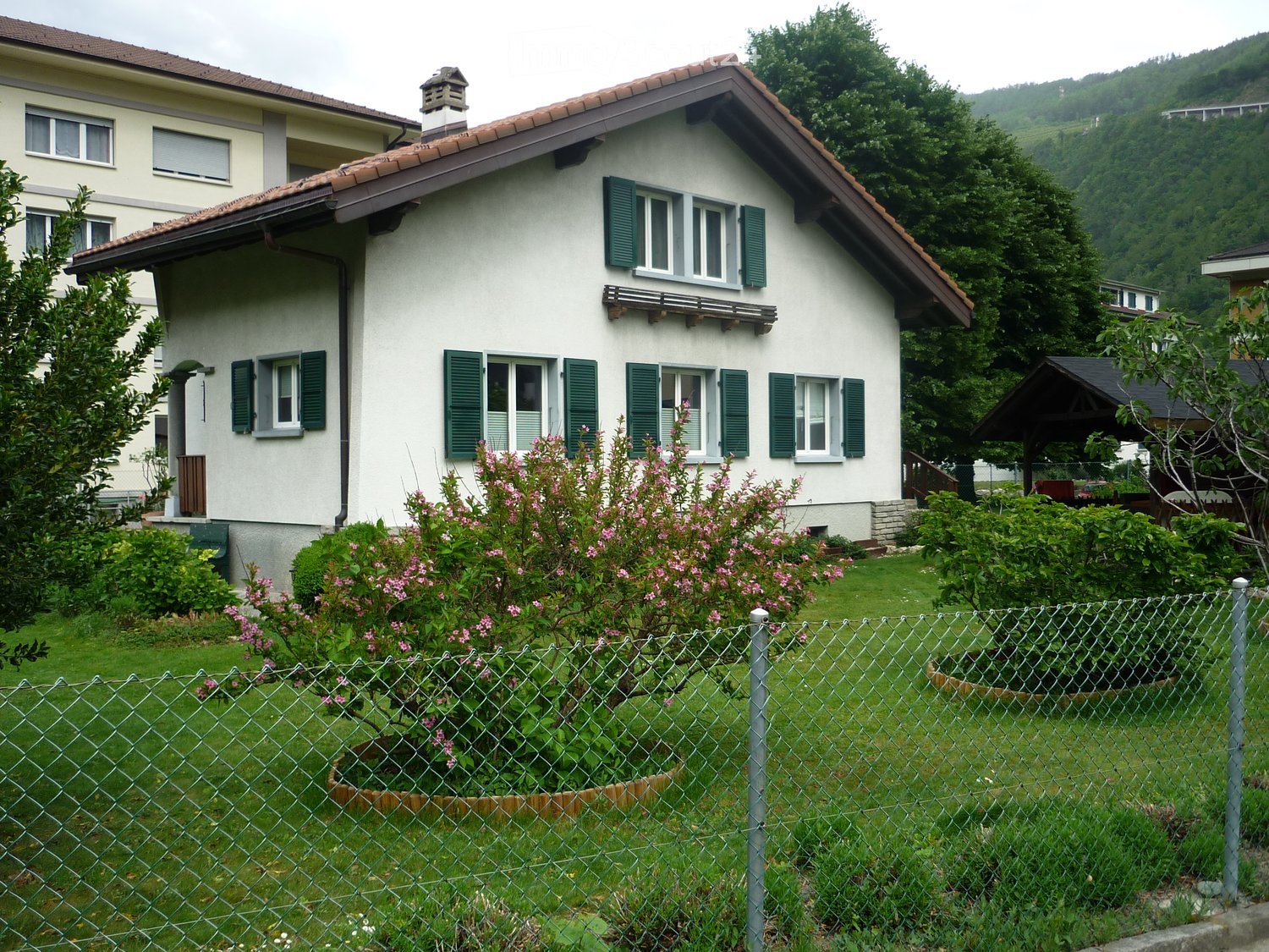 Two-story house with white walls, green shutters, and a red-tiled roof, surrounded by a well-maintained garden with flowering shrubs and trees.
