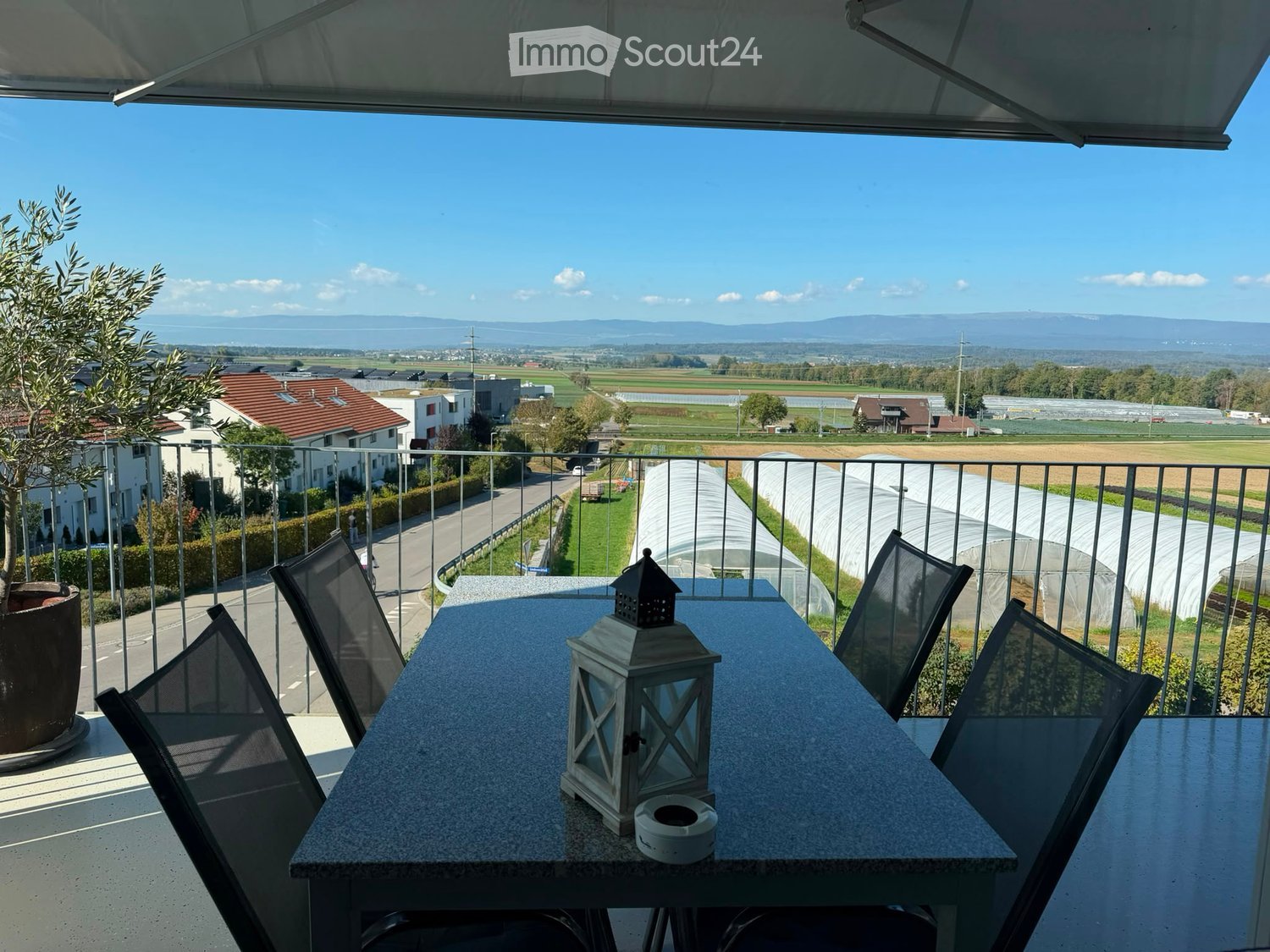 Balcony with a table, chairs, and a lantern, overlooking a rural landscape with fields and houses.