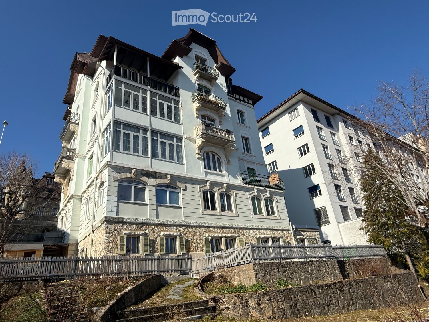 A multi-story residential building with light-colored walls, multiple windows, and balconies, situated on a raised ground floor with stone steps leading up to it.