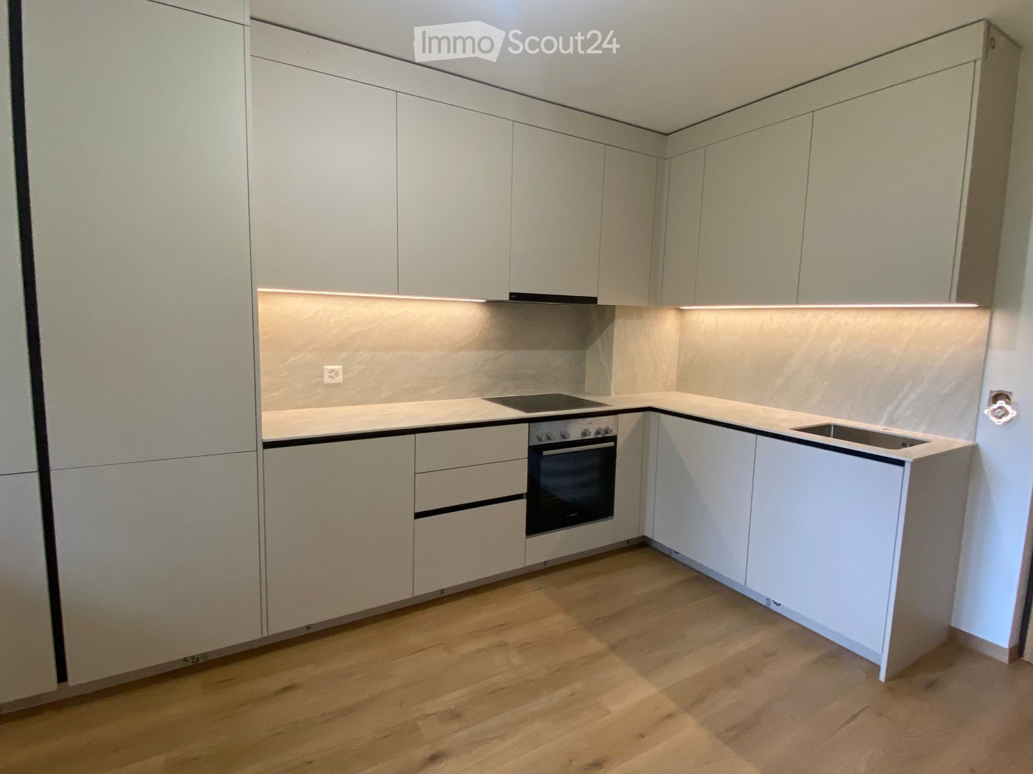 Modern kitchen with light grey cabinets, wooden flooring, and built-in appliances including an oven, induction cooktop, and sink.
