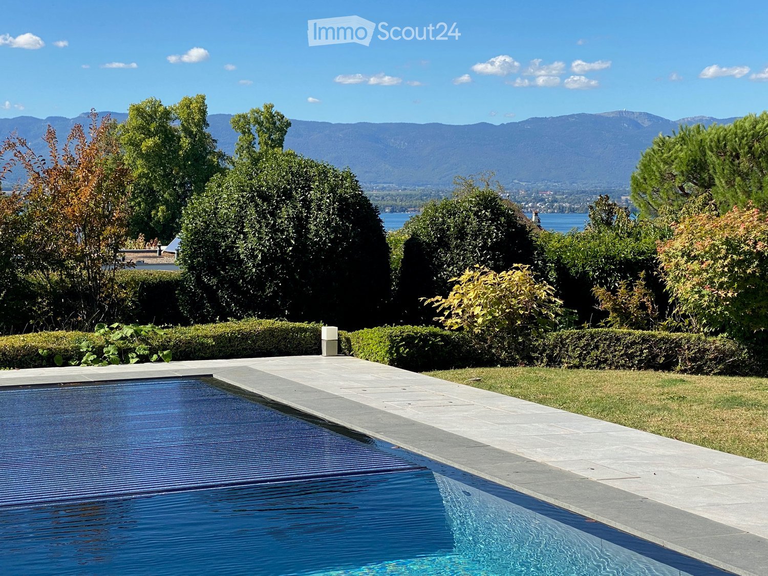 A well-maintained garden with a swimming pool, surrounded by neatly trimmed hedges and lush greenery, with a scenic mountain and lake view in the background.