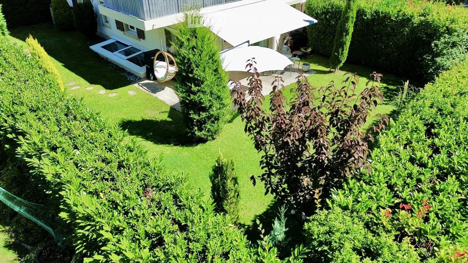 Well-maintained garden with green lawn, various shrubs, a hanging egg chair, patio umbrella, and a covered outdoor seating area.
