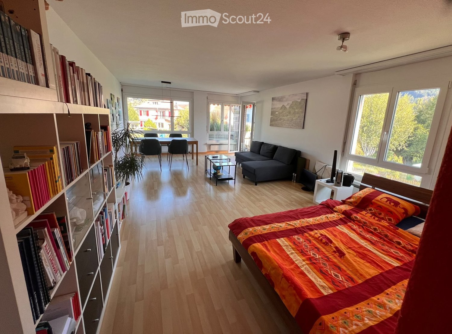 A well-lit, open-plan living and sleeping area with a bed, sofa, dining table, bookshelf, and large windows offering a view of the outdoors.