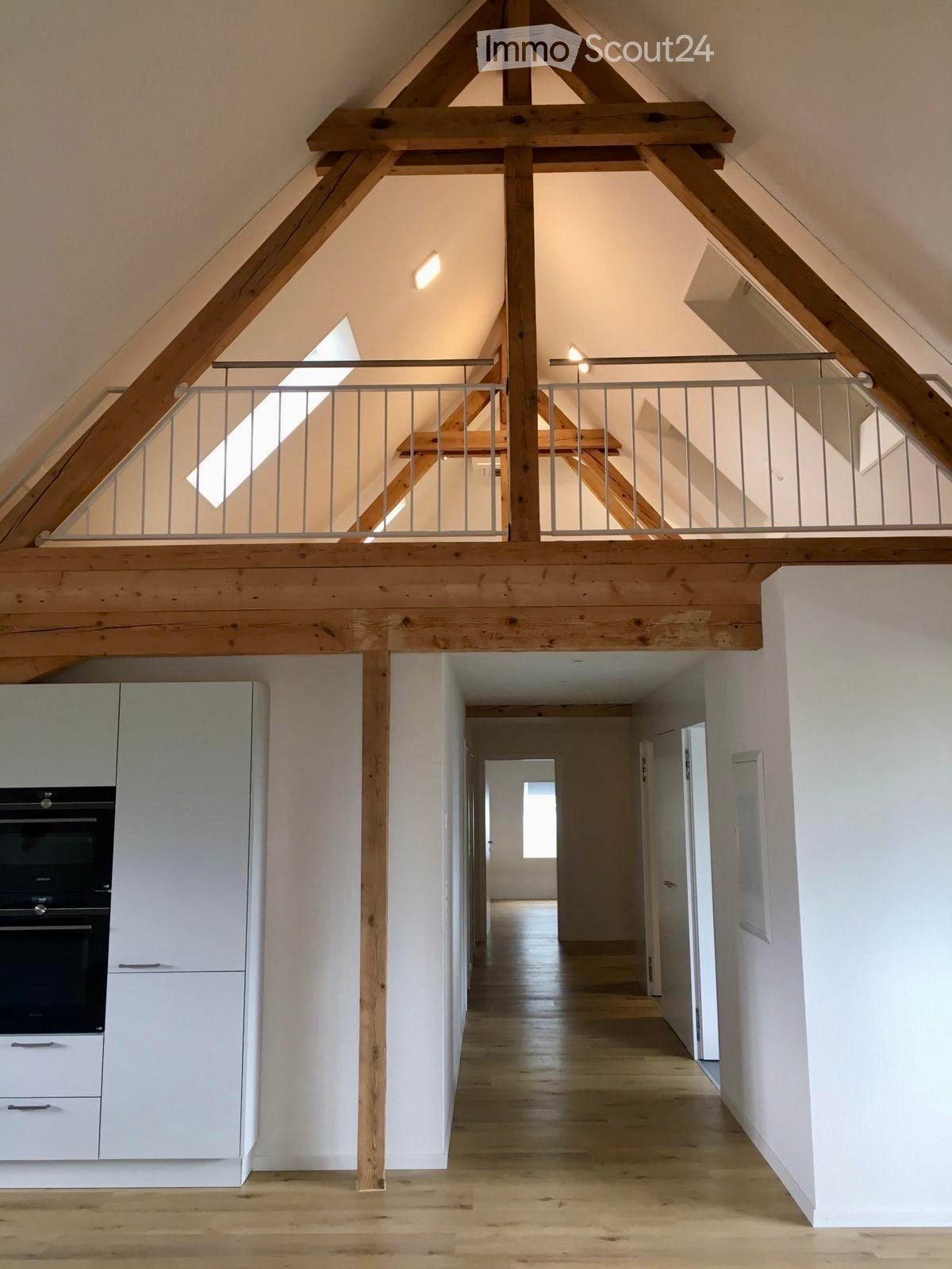 Bright, open hallway with wooden floors and white walls, leading to a kitchen area with white cabinets and a built-in oven. The hallway features exposed wooden beams and a metal railing leading to an upper level.
