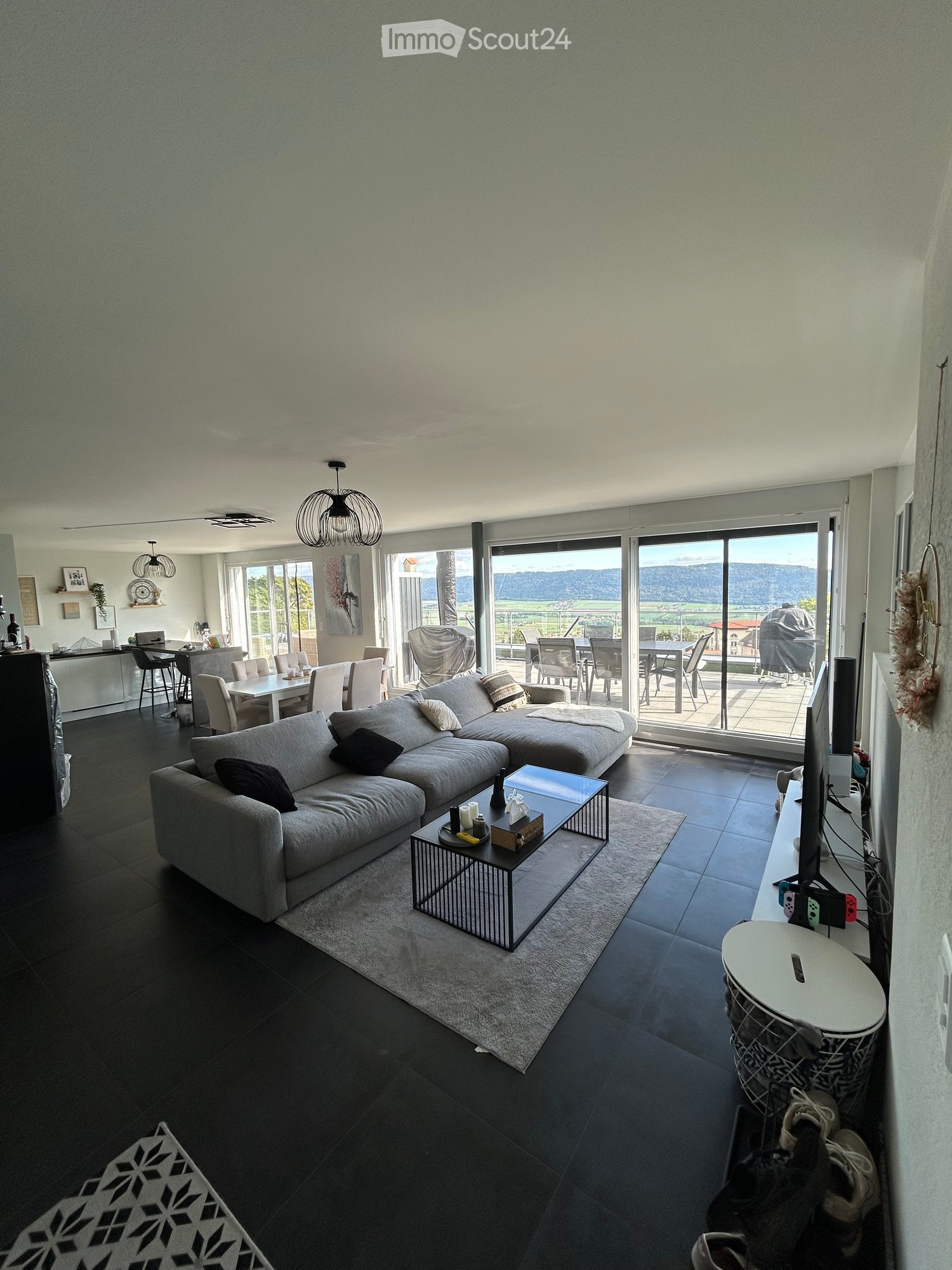 Modern living room with dark tiled flooring, grey sectional sofa, coffee table, dining area with chairs and table, and a view through large windows.
