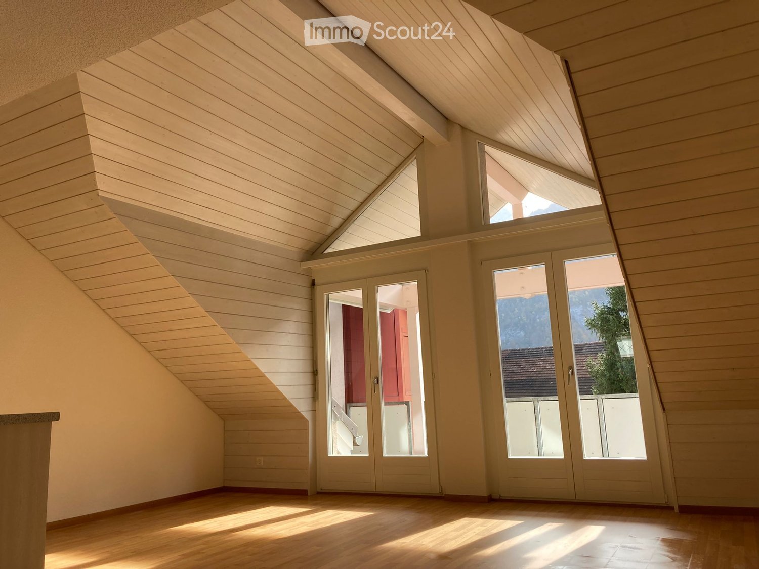 Bright, empty attic room with wooden walls, slanted wooden ceiling, large windows, and double doors leading outside.
