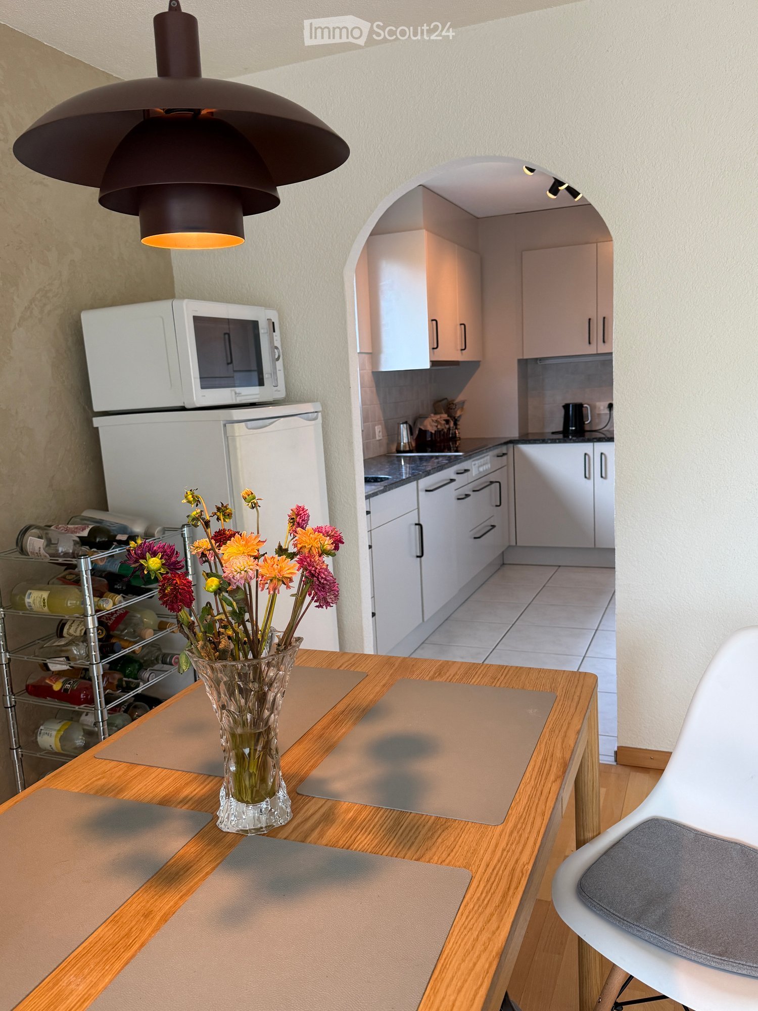 Modern kitchen with white cabinets, wooden table, vase with flowers, microwave, refrigerator, and wine rack.