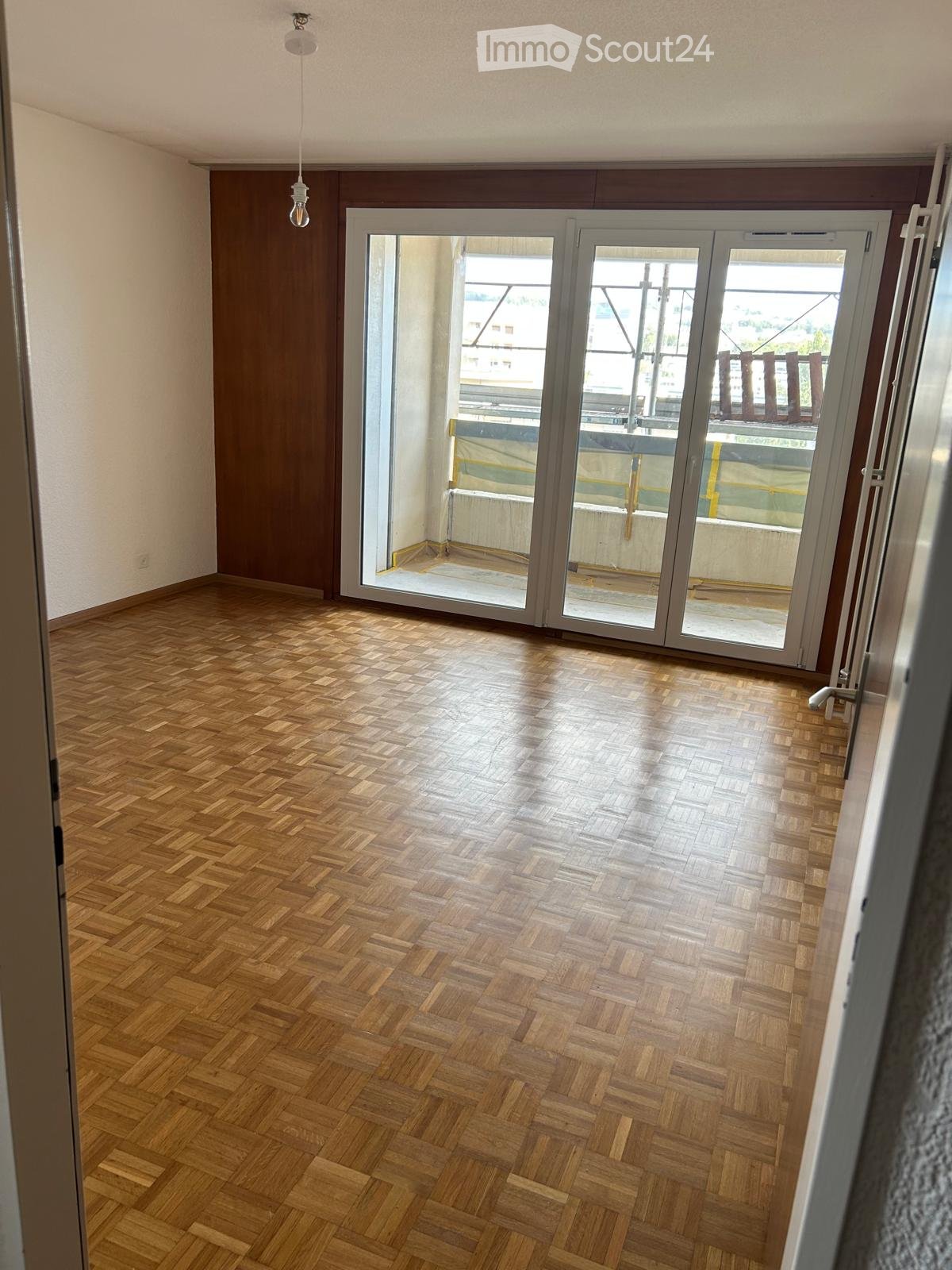 Empty room with wooden herringbone flooring, large sliding glass doors leading to a balcony, and a visible light fixture on the ceiling.
