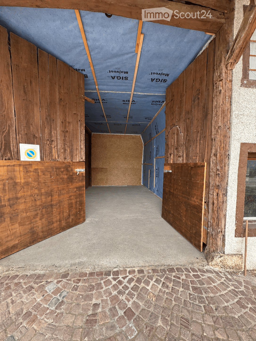 Empty corridor with exposed wooden beams, concrete floor, and cobblestone pathway. Wooden doors on both sides, with insulation visible in the ceiling area.
