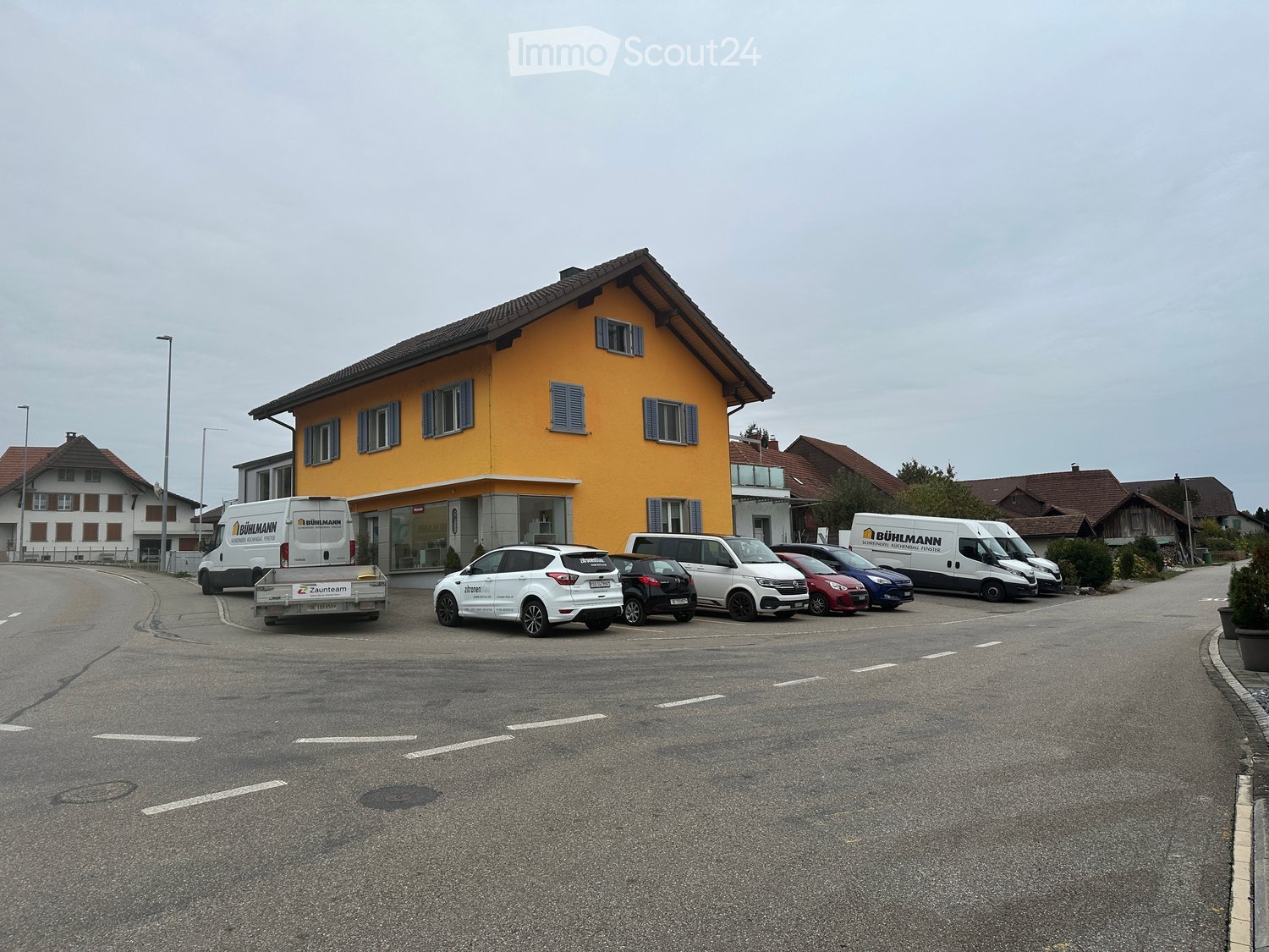 Yellow two-story building with multiple windows and shutters, surrounded by parked cars and vans in a lot.