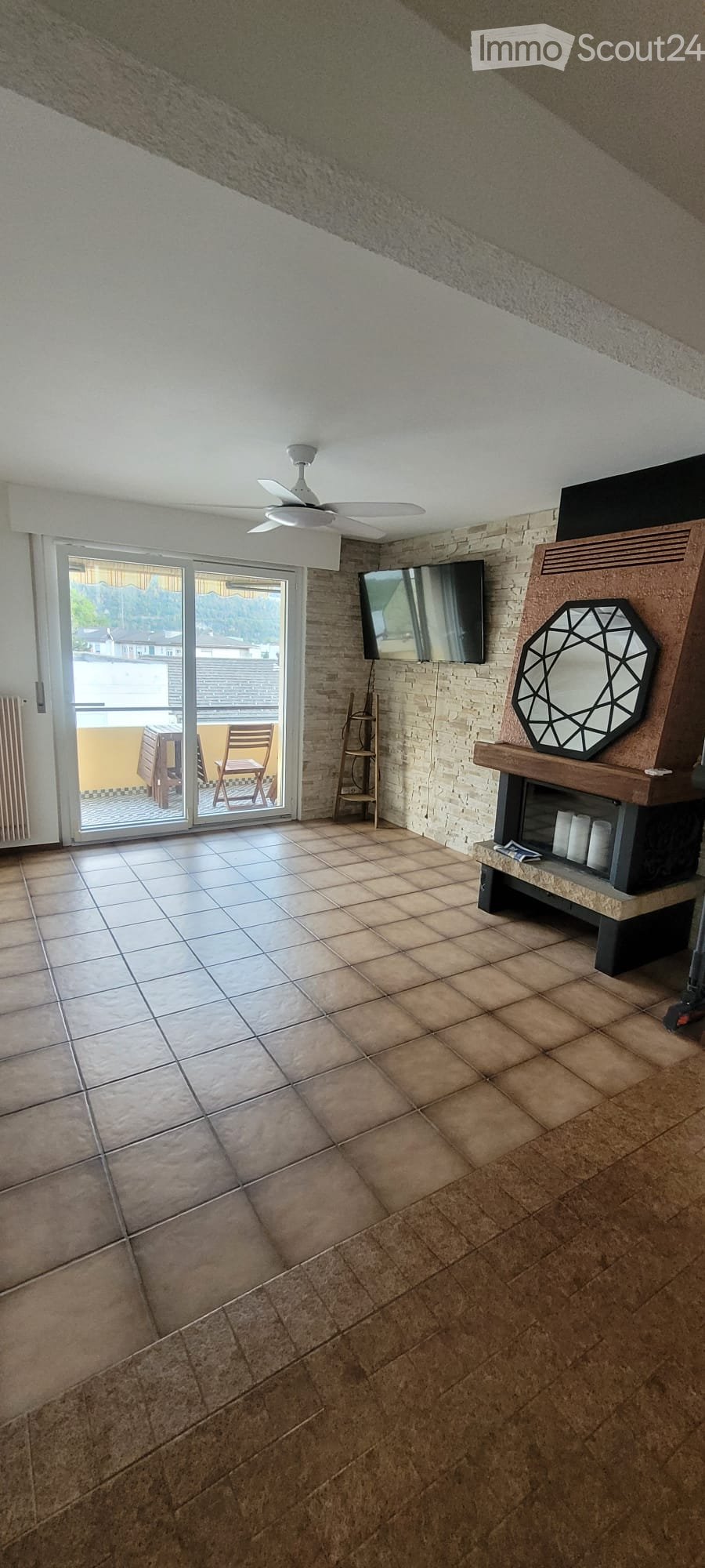 Living room with tiled floor, white ceiling fan, television mounted on wall, decorative mirror on wooden stand, and sliding glass door leading to balcony with outdoor furniture.