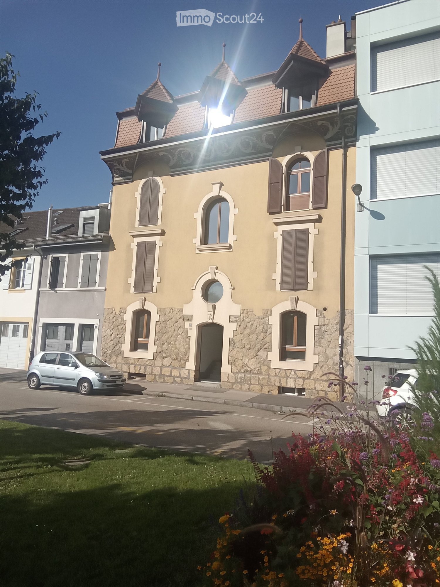 A two-story house with a beige facade, arched windows, and a decorative roofline. The house features stone detailing around the base and has multiple windows with shutters. There is a small garden with colorful flowers in the foreground, and a parked car
