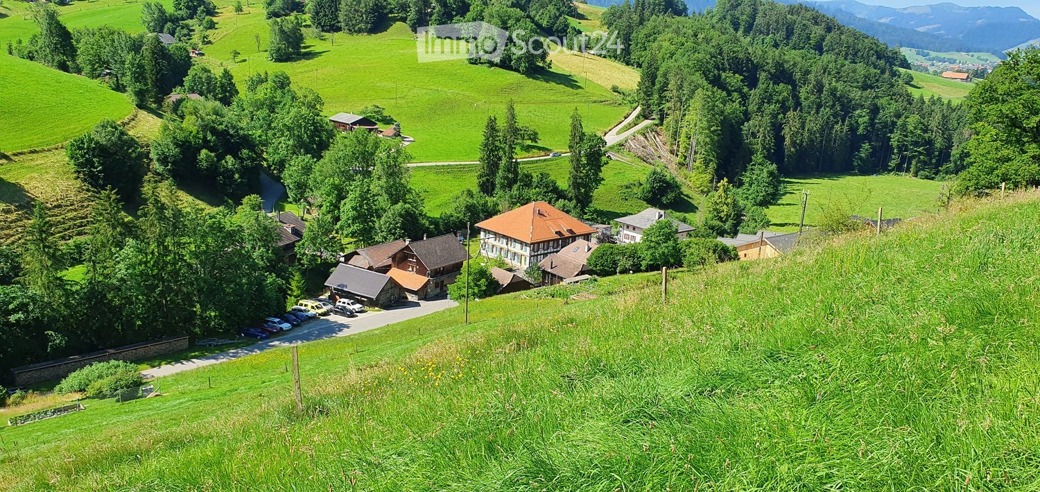 A cluster of houses and buildings in a lush green valley with rolling hills and trees, surrounded by a parking area.