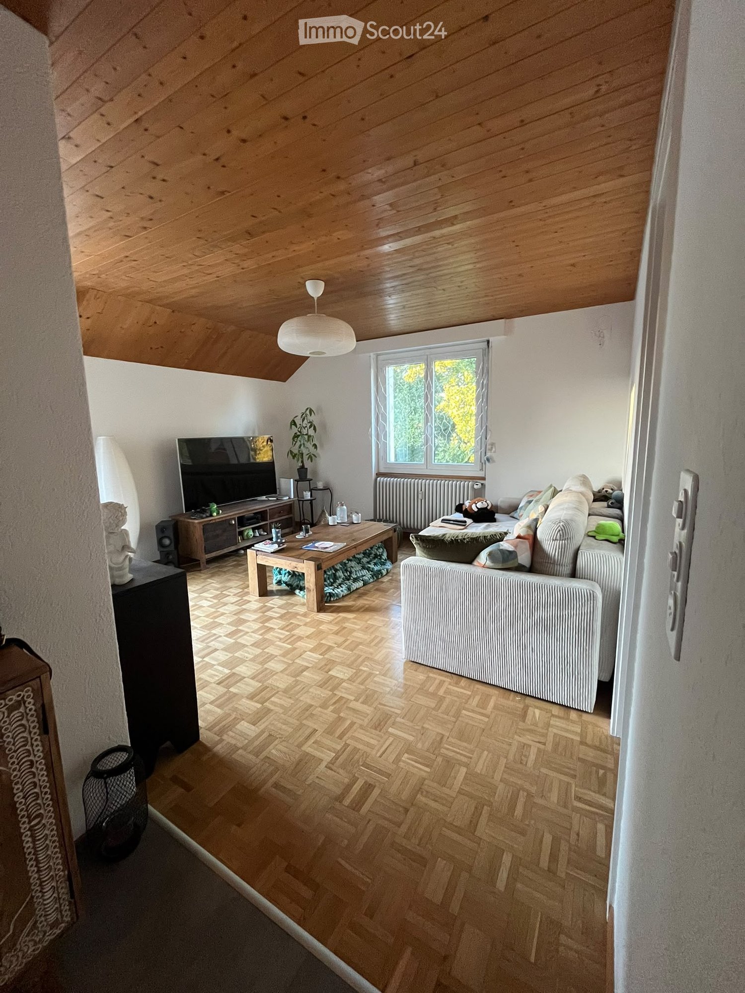 Living room with wooden herringbone flooring, white walls, exposed wooden ceiling beams, large window with view, sofa, coffee table, TV, plant, floor lamp, radiator, side table, speaker.