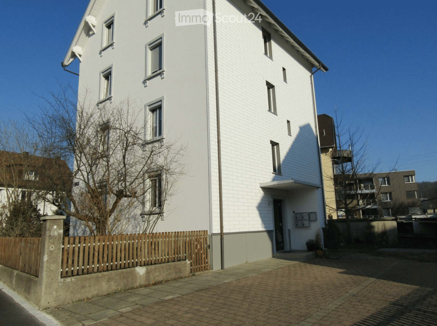 A modern, three-story white house with multiple windows, a wooden fence, and a small garden area.