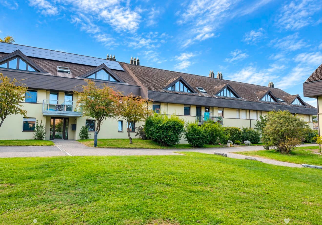 Two-story residential building with a modern design, featuring a sloped roof, large windows, and solar panels on the roof. The building is surrounded by a well-maintained lawn and trees.