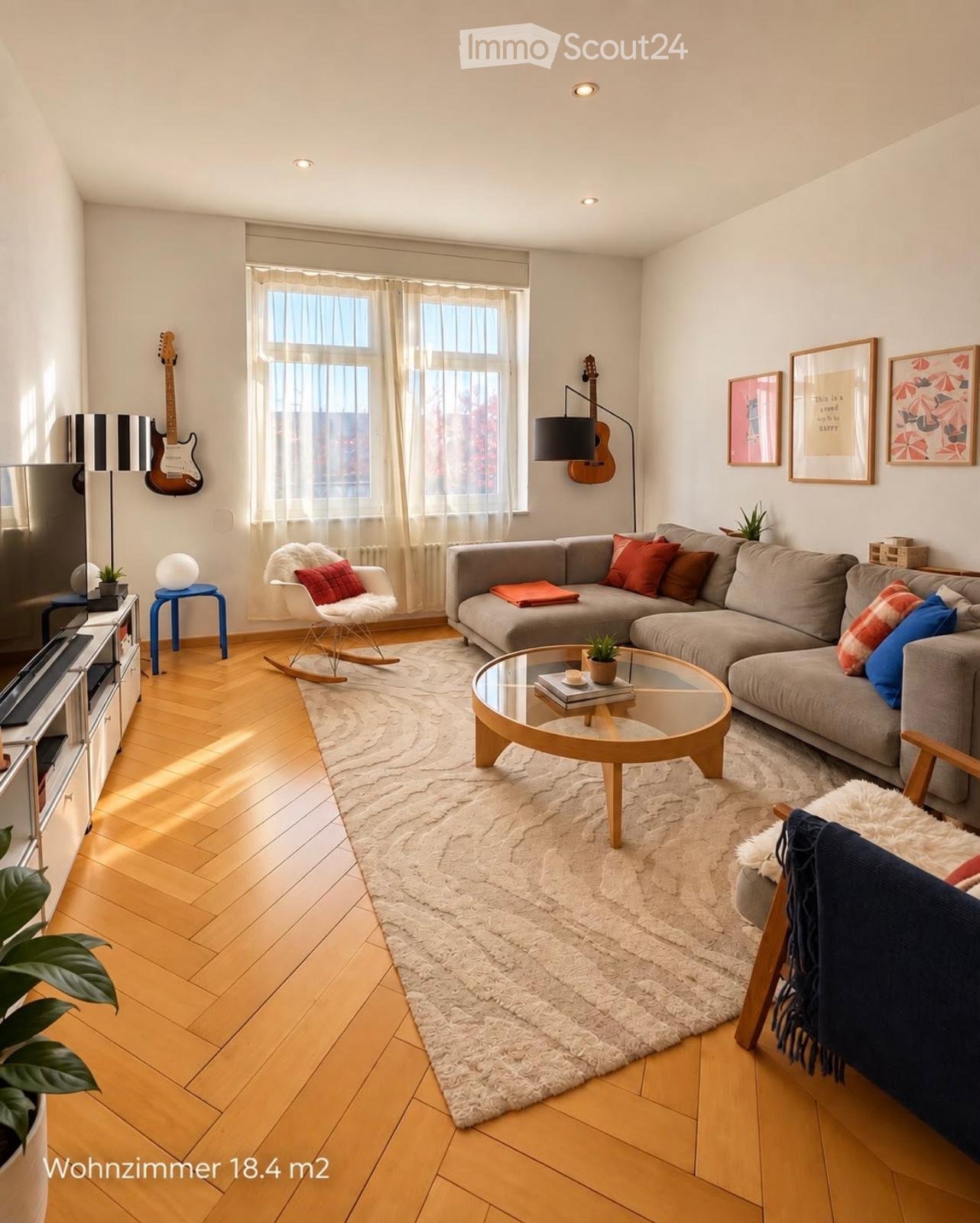 Modern living room with wooden flooring, large window with sheer curtains, grey sectional sofa, glass coffee table, rocking chair, guitar on wall, TV stand, and decorative items.