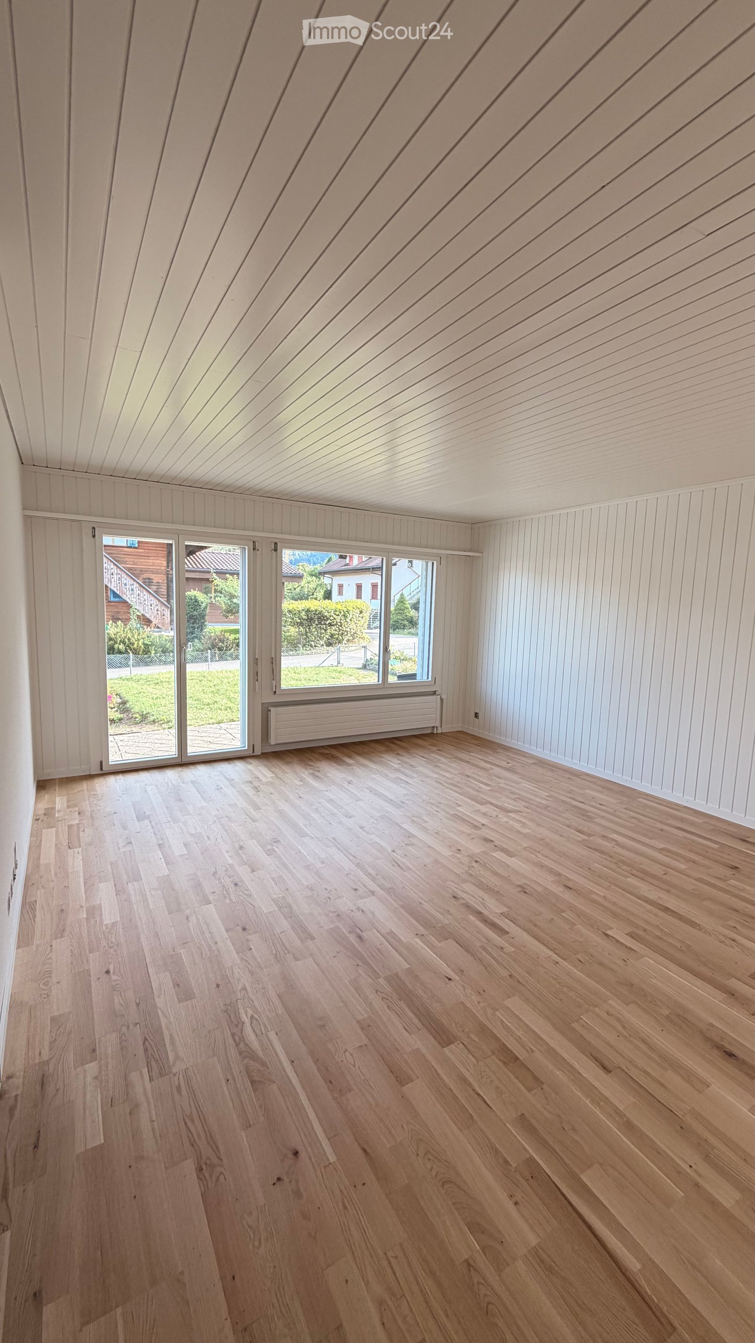 Empty room with light wooden flooring, white paneled walls, and a slanted wooden ceiling. Large windows and sliding doors lead to an outdoor area.