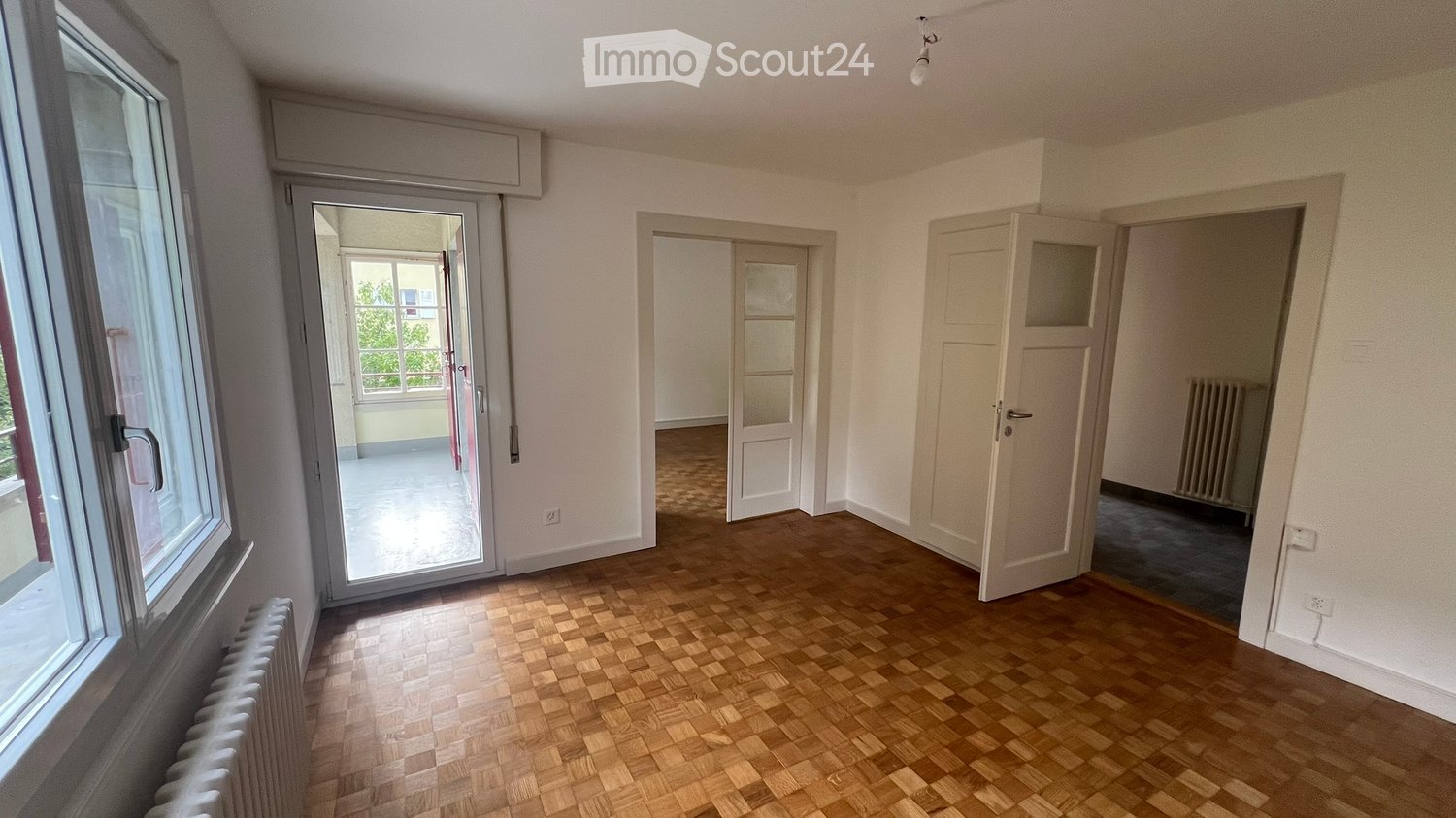 Empty room with wooden flooring, white walls, and multiple doors leading to other spaces. Natural light from windows illuminates the space.