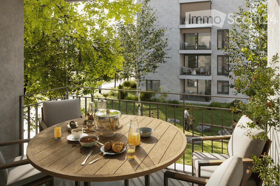 Balcony with a round wooden table set for breakfast, surrounded by chairs and a railing, overlooking a green park and modern apartment building.