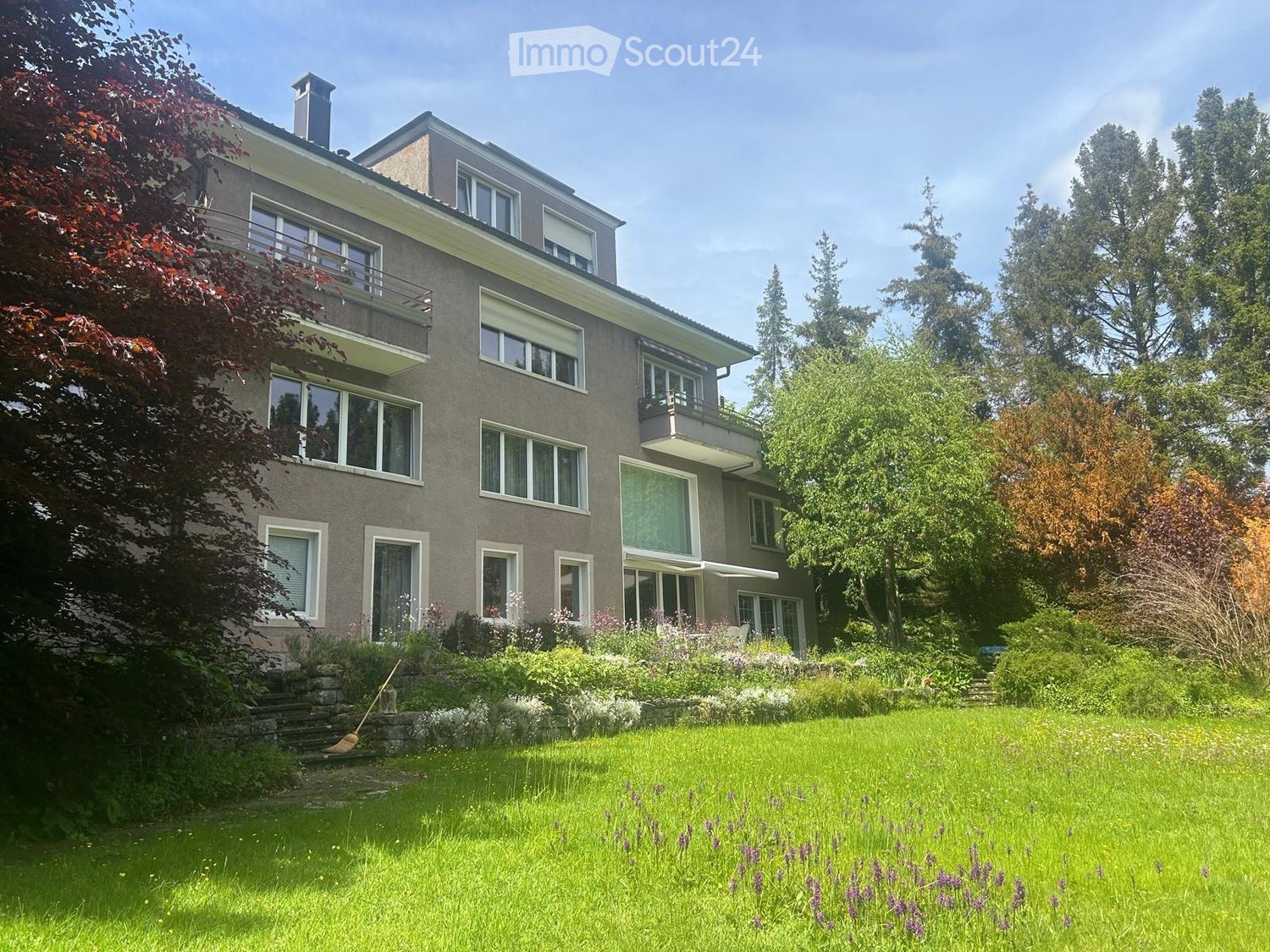 Two-story house with grey facade, multiple windows, balconies, and a well-maintained garden with green lawn and flowering plants.