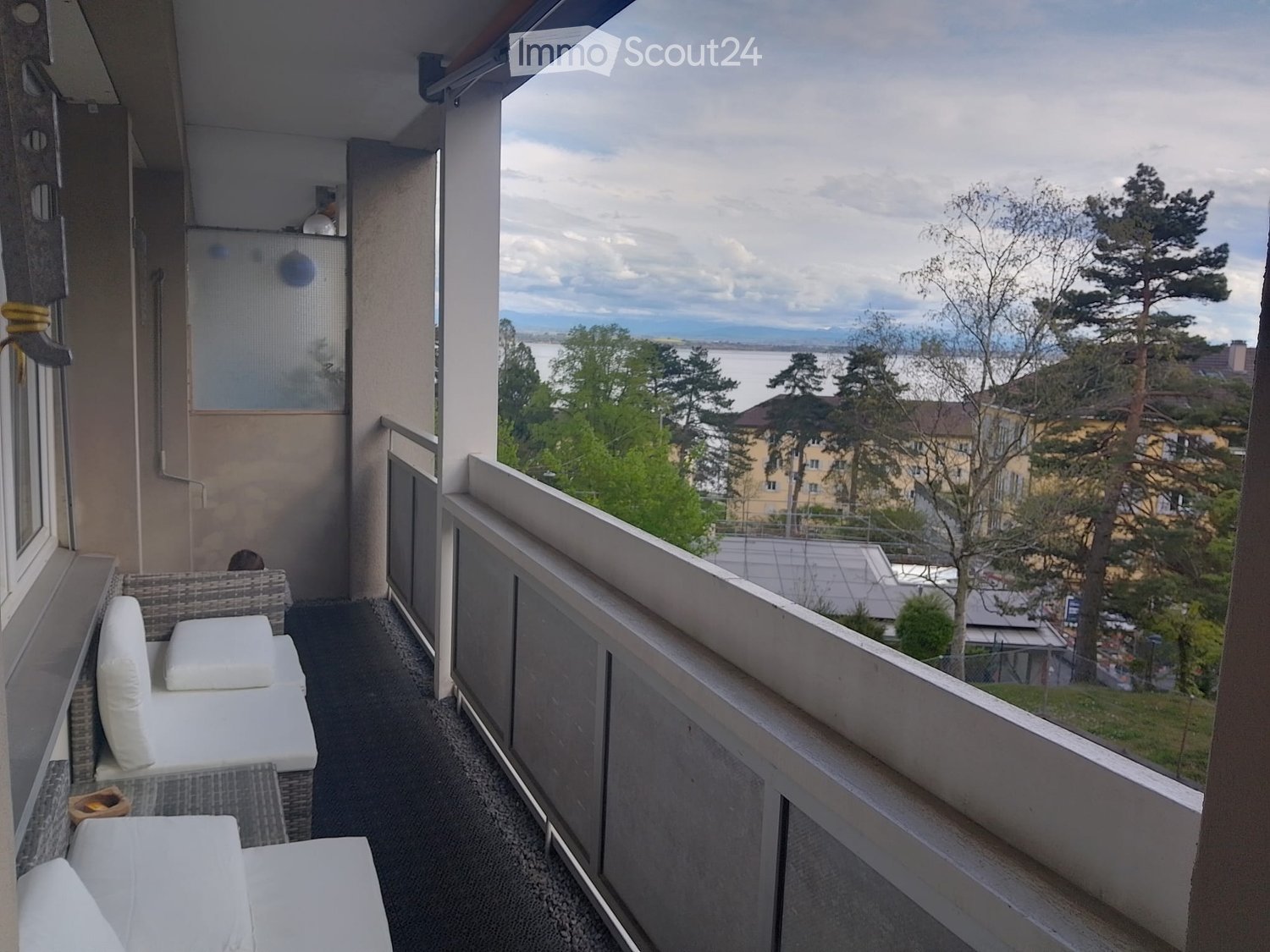 Balcony with white cushions, a small table, and a scenic view of a lake and surrounding trees.