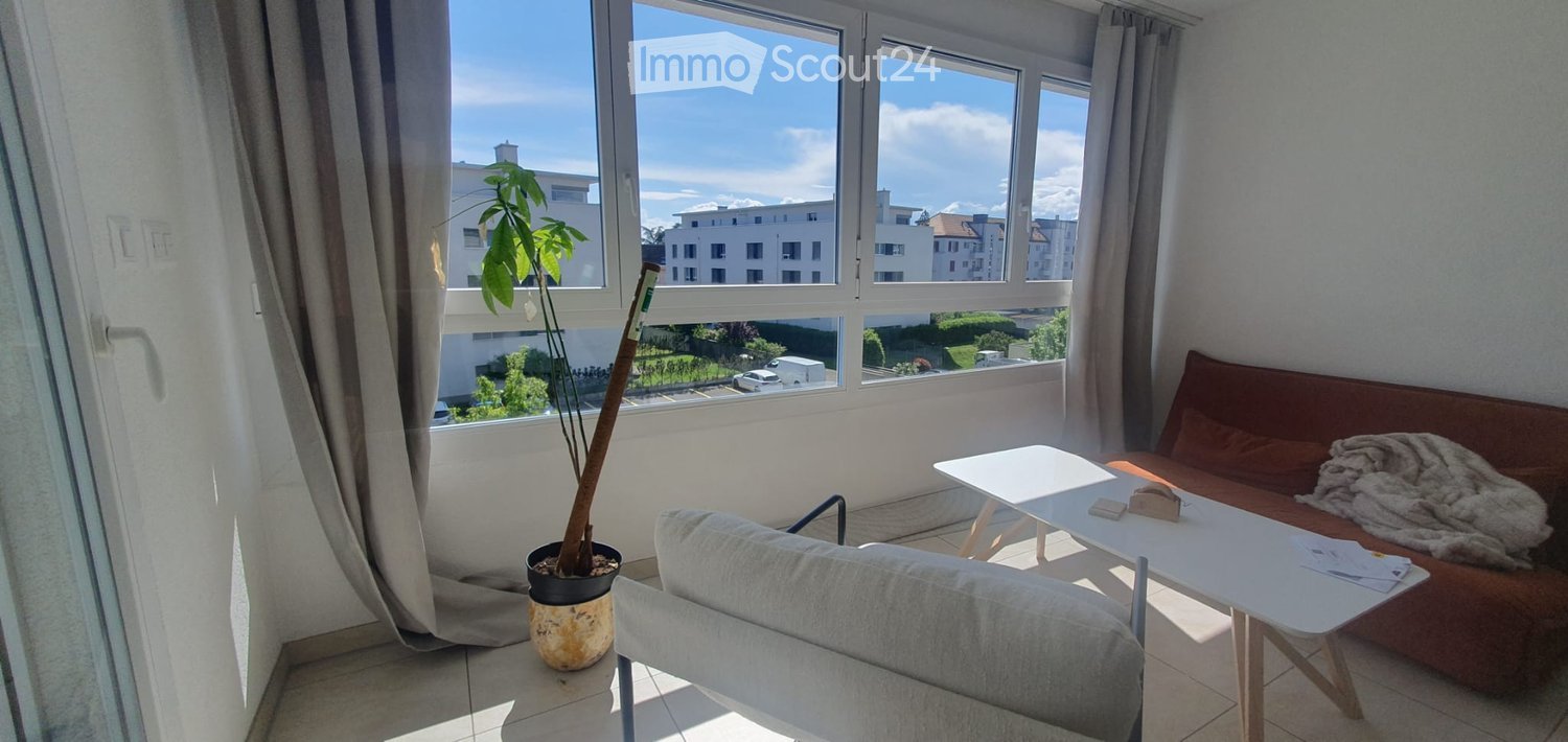Bright living room with large windows offering a view of the cityscape, featuring a grey sofa, white coffee table, armchair, and a potted plant.