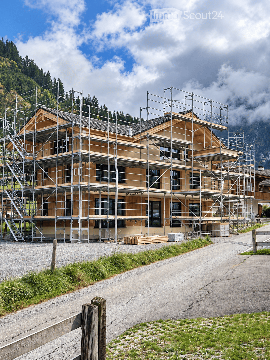 A two-story house under construction, covered in scaffolding, with wooden framing and large windows, situated in a mountainous area with a gravel driveway and surrounding greenery.