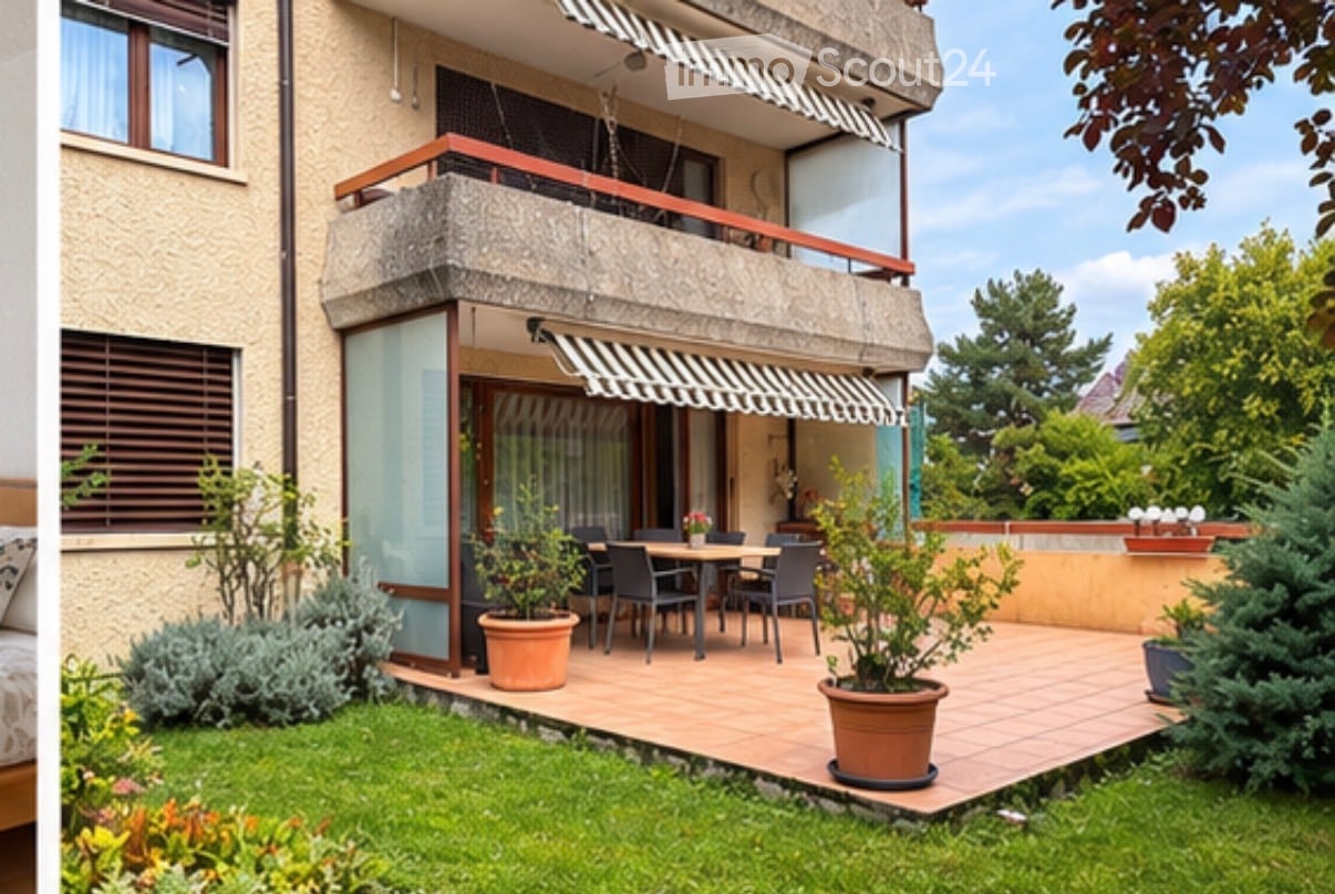 Balcony with a dining table and chairs, surrounded by potted plants and greenery, attached to a multi-story building with striped awnings.