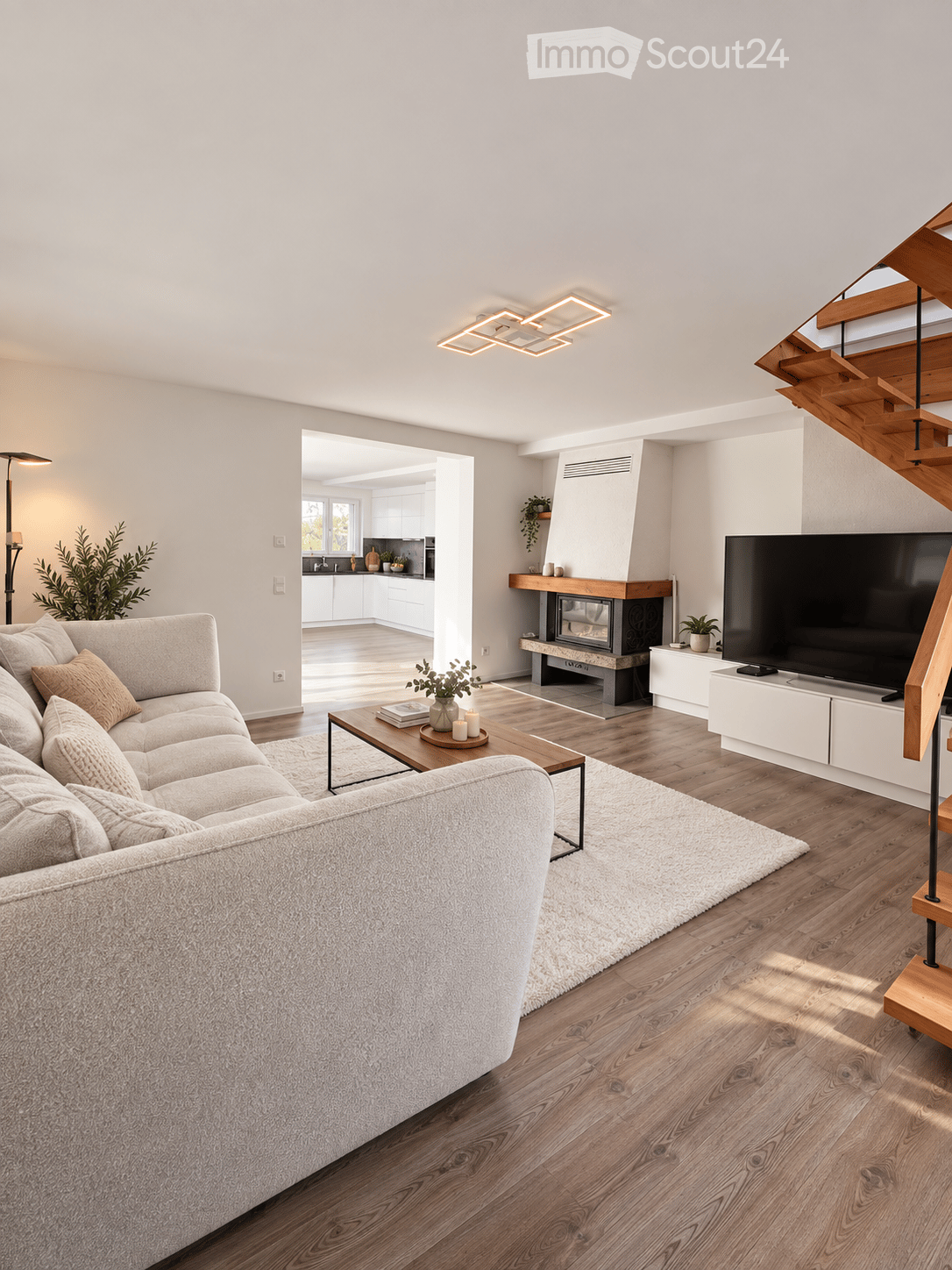 Modern living room with a large sectional sofa, coffee table, fireplace, and television. The space features wooden flooring, recessed lighting, and a view into an adjacent kitchen area.