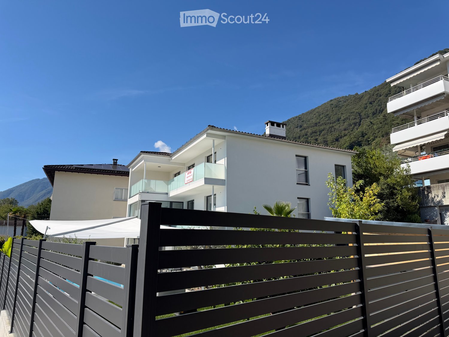 Modern two-story white building with balconies and large windows, surrounded by greenery and a mountainous backdrop.