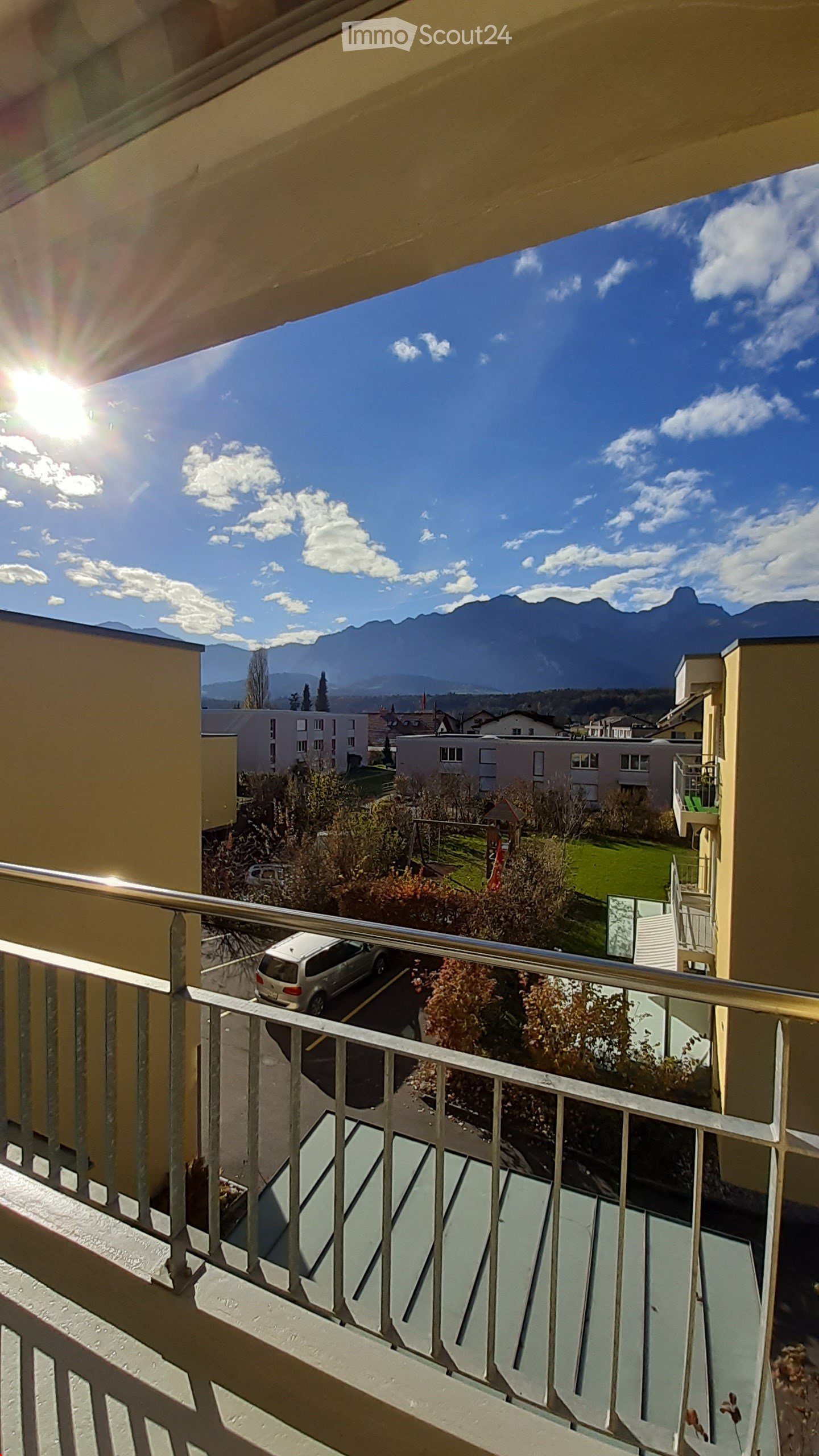 Balcony with metal railing, overlooking a residential area with buildings, a playground, and distant mountains under a clear blue sky.