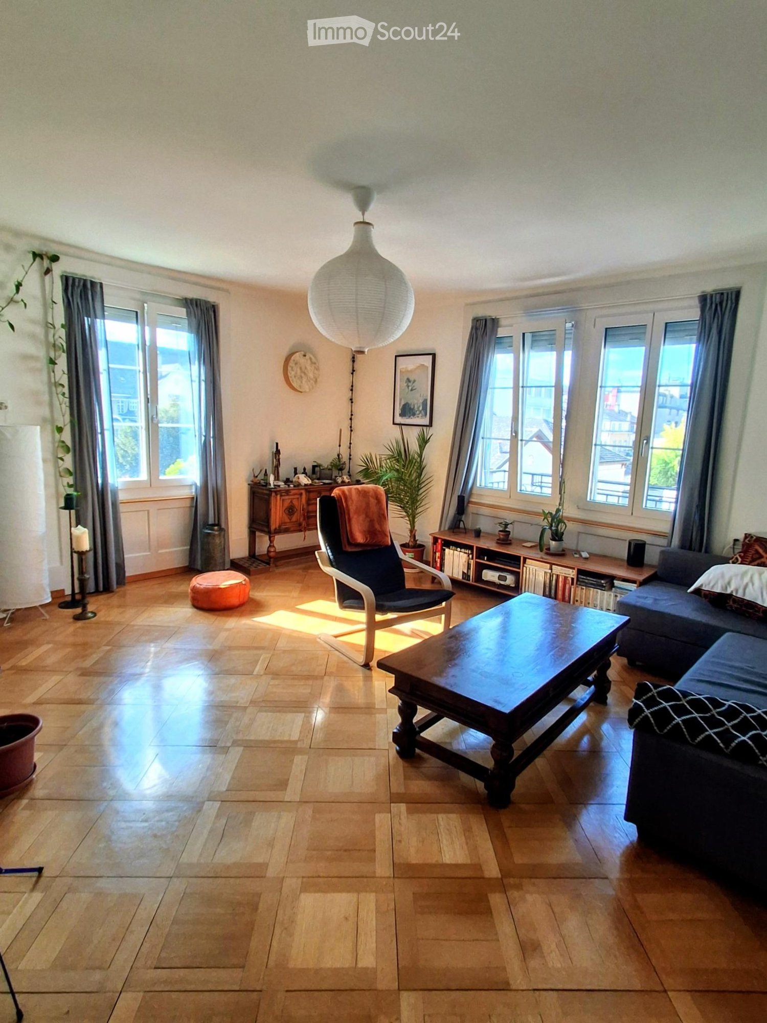 Bright living room with wooden flooring, large windows, a sofa, coffee table, armchair, and decorative plants.
