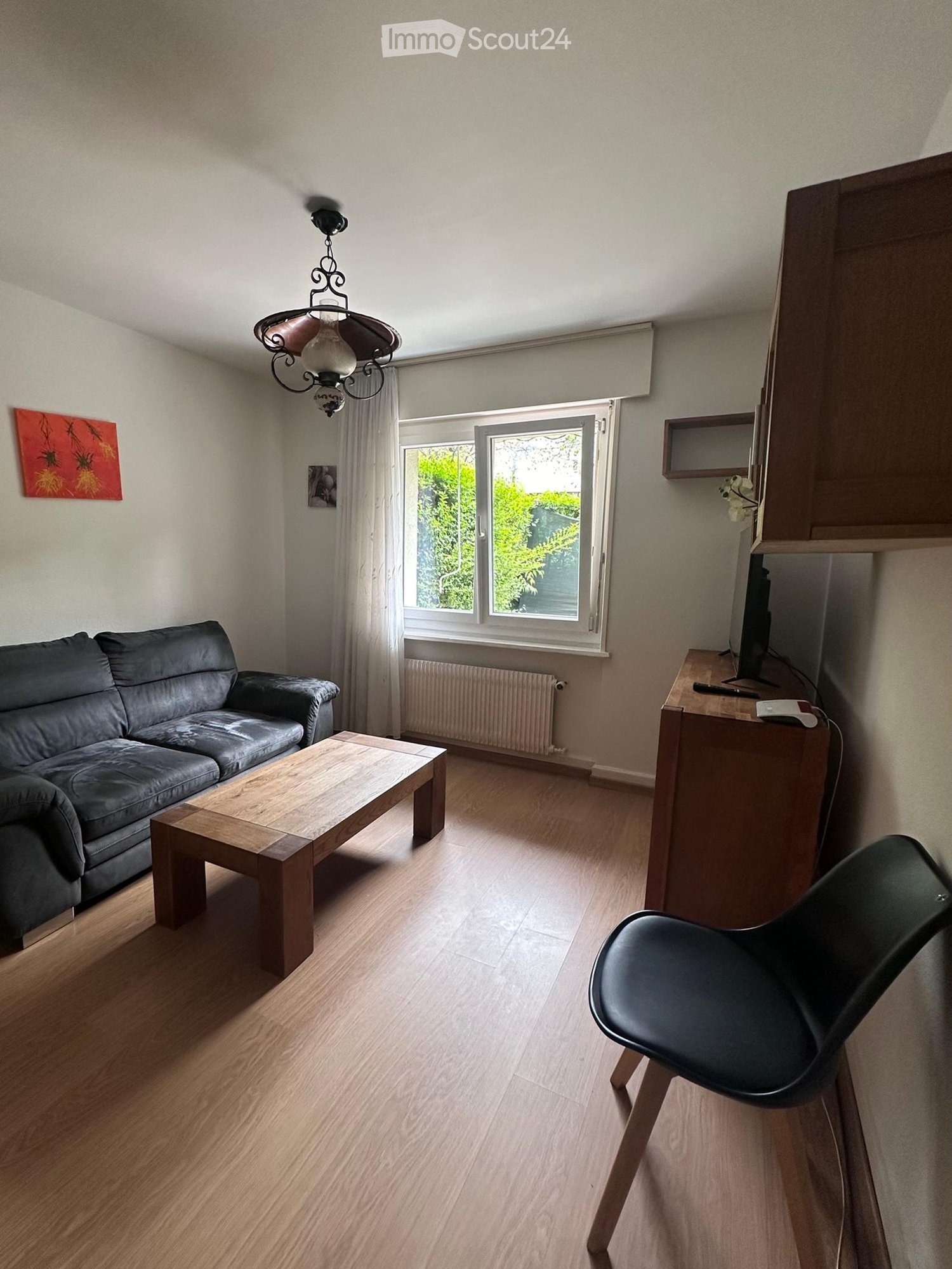 Living room with wooden flooring, large window with white curtains, grey sofa, wooden coffee table, black chair, TV stand, and a decorative painting on the wall.
