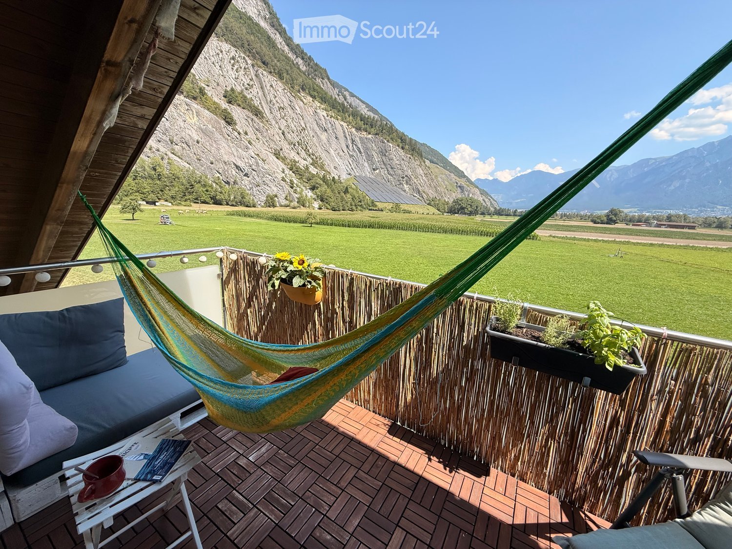 Balcony with wooden decking, bamboo privacy screen, hammock, sofa, small table with a cup, potted plants, and a scenic mountain view.