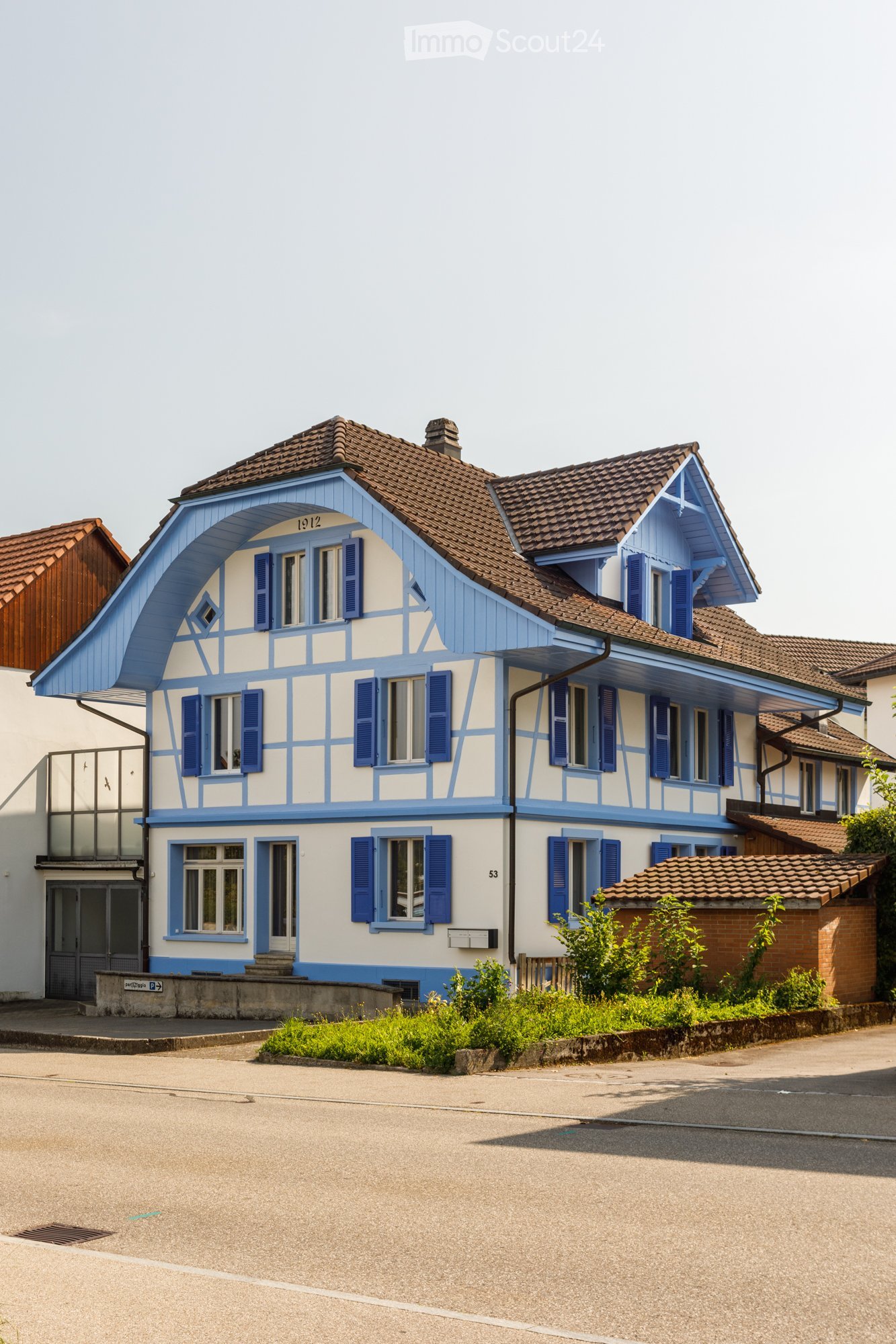 Two-story house with blue and white exterior, brown tiled roof, multiple windows with shutters, and small garden area.