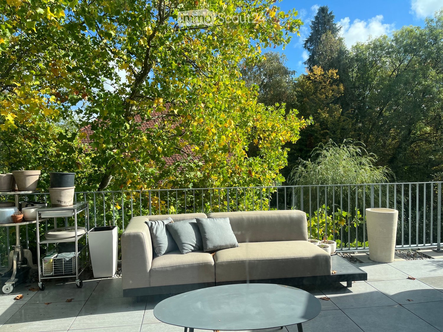 Balcony with a modern sofa, coffee table, side table, and potted plants, surrounded by trees and a clear blue sky.