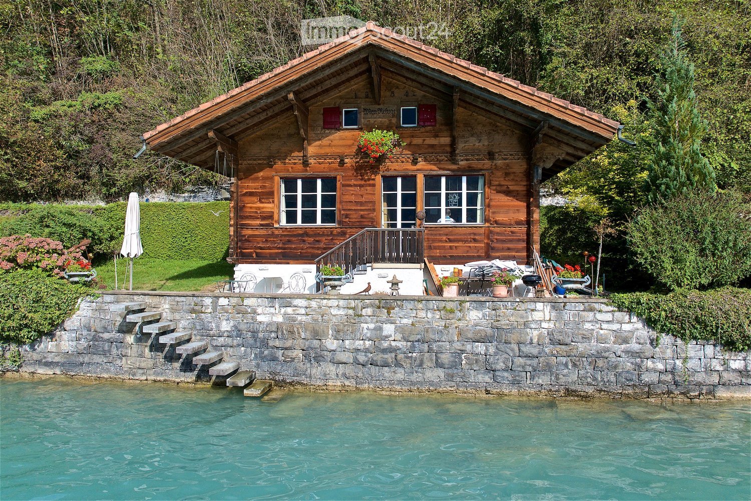 A wooden house with a sloped roof, large windows, and a balcony overlooking a swimming pool. The house is surrounded by greenery and features a stone wall and steps leading to the pool.
