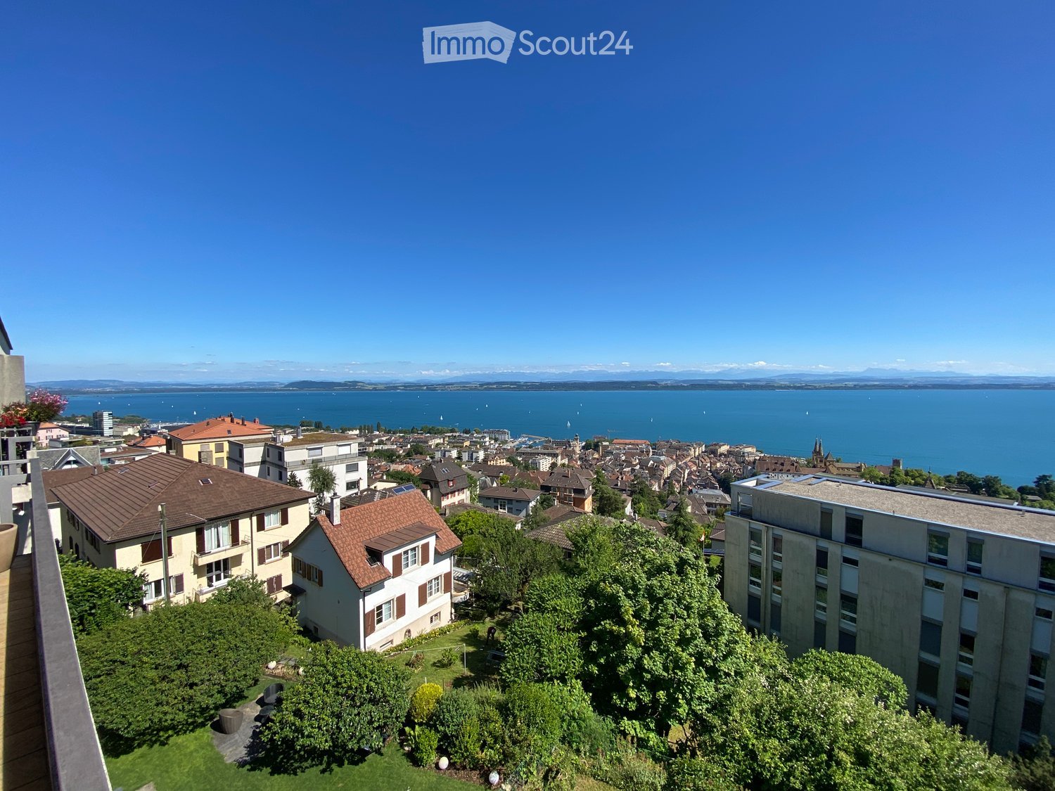 A panoramic view of a town with houses, trees, and a large body of water in the background under a clear blue sky.