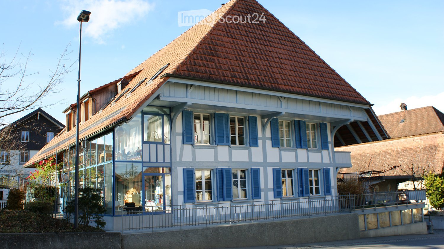 Two-story house with a red-tiled roof, white and blue exterior, large windows, and a small balcony area.