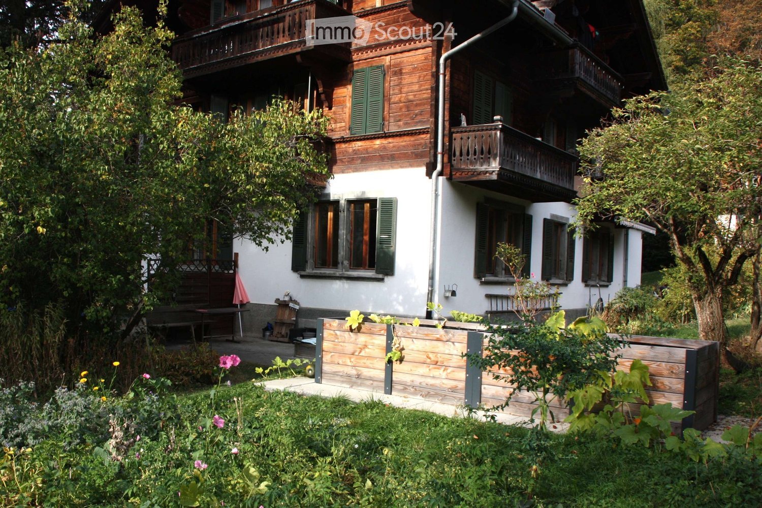 A two-story wooden house with green shutters, balconies, and a well-maintained garden with flowers and a wooden planter box.